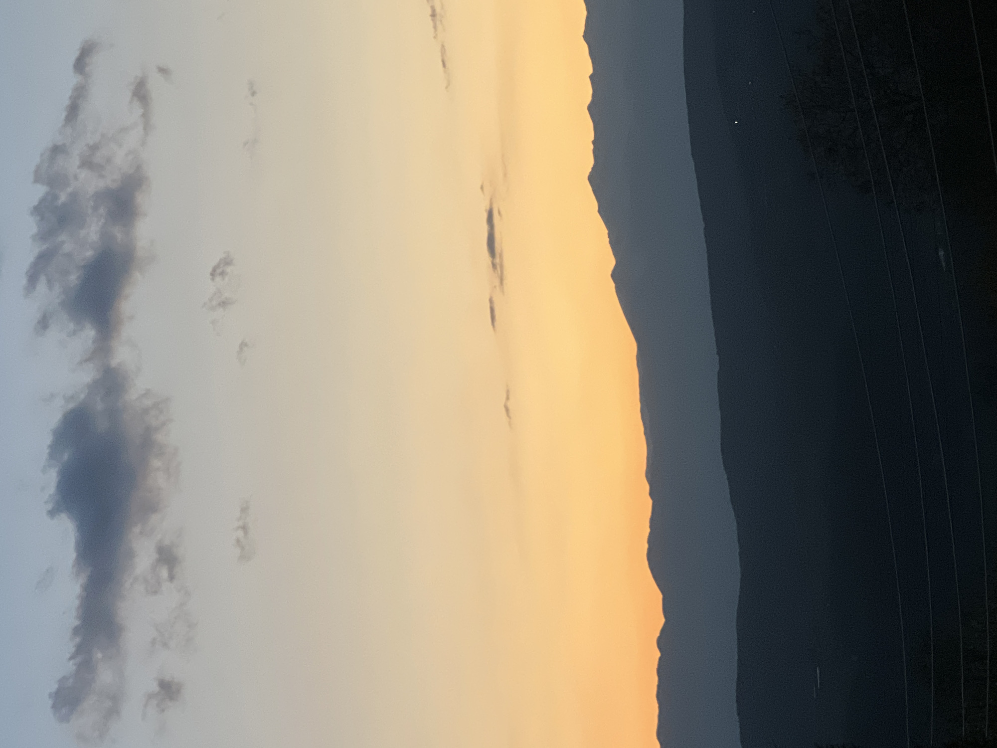 The image shows a sunset in Kribul, Bulgaria, with a mountain range silhouetted against the orange and yellow sky. There are a few clouds in the sky, and some power lines are visible in the lower right corner.