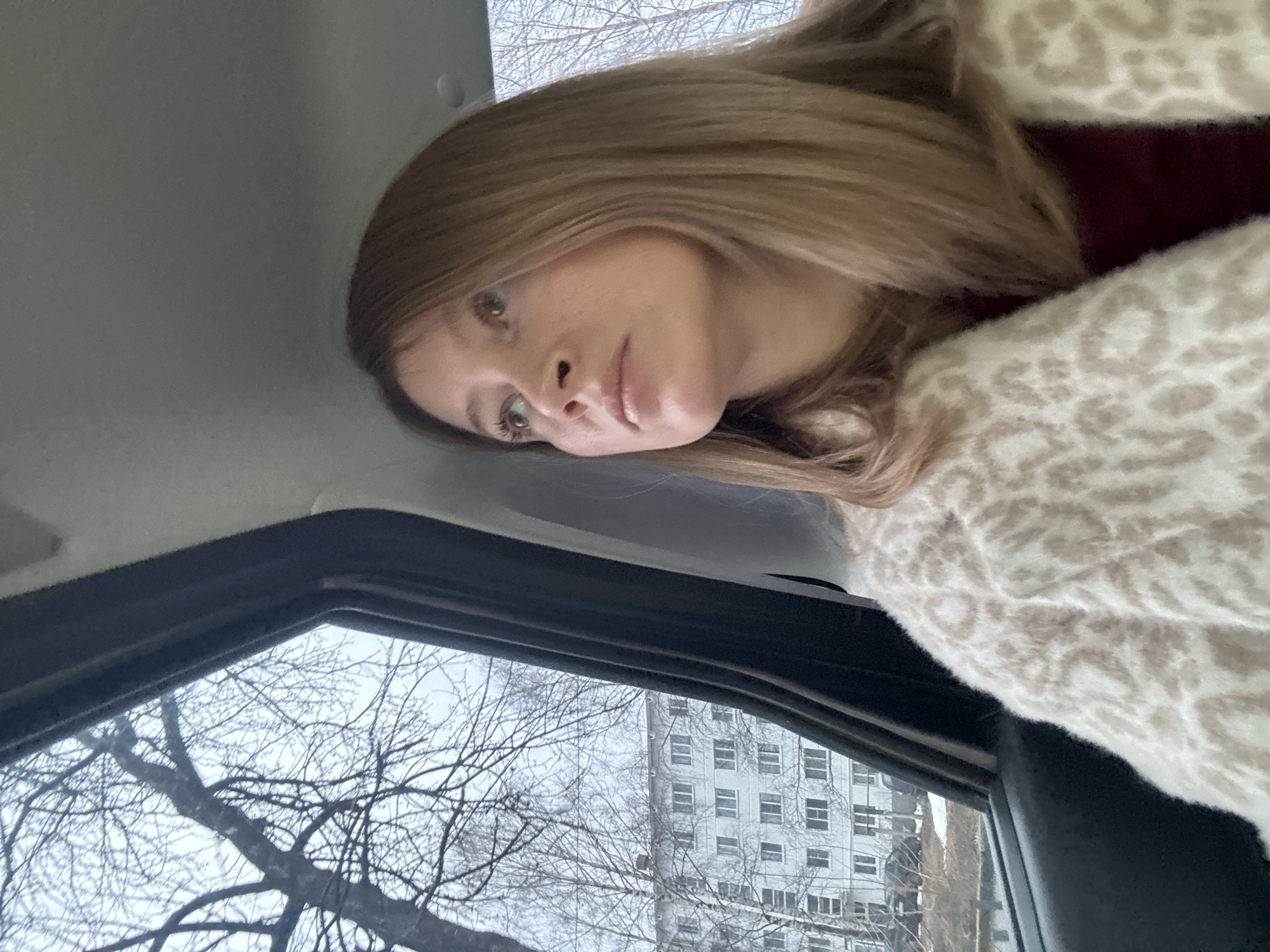 A woman with light brown hair is inside a vehicle. She is wearing a patterned, possibly leopard-print, fuzzy cardigan over a maroon top. The view through the car window shows bare trees and a building with multiple windows. Based on the architectural style of the building visible through the window, and the woman's potential ethnicity, it is likely that this image was taken in Russia. Without further information, a specific location within the city of Chaikovsky cannot be confirmed.