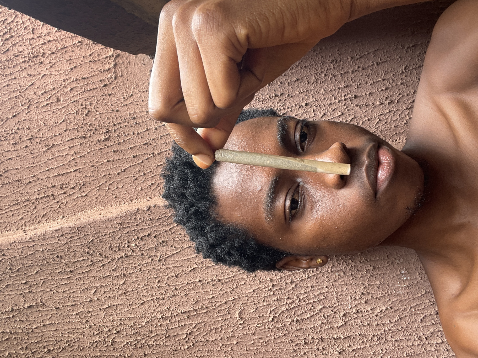 A person is lying on a rough, textured surface, possibly concrete, indoors. They are holding a hand-rolled cigarette or joint between their fingers, positioned over their upper lip and nostrils. The person has short, dark curly hair, and their skin tone is brown. The location is the city of Nkpor, Nigeria.
