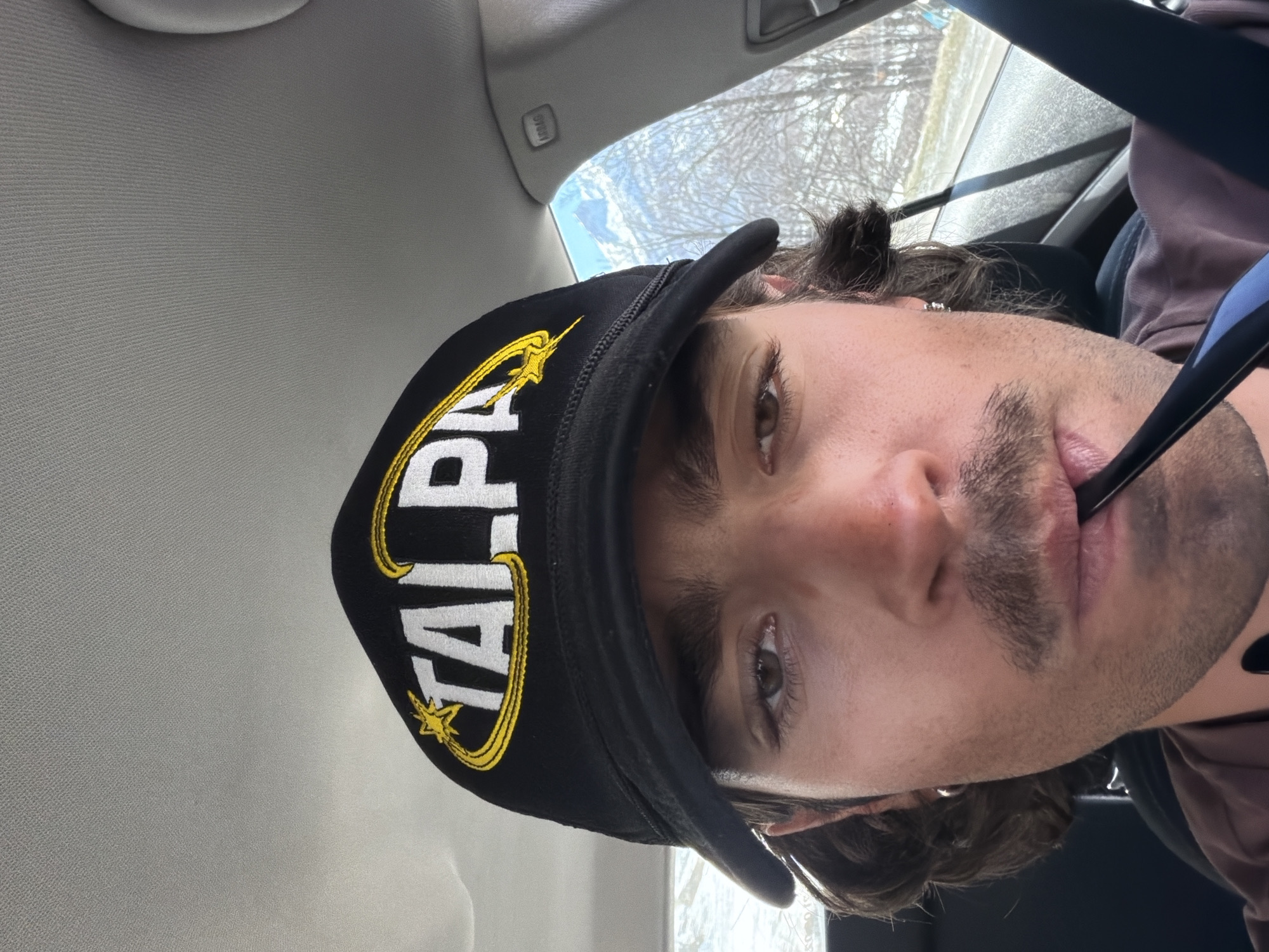 A man is wearing a black cap with white "TALPA" lettering and a yellow star/crescent design, positioned inside a car. The man is fair-skinned, with dark brown hair, light brown eyes, and a mustache/goatee. A blue object is in his mouth. The car's interior is visible, including the ceiling and airbags. Outside the window, trees and snow-covered ground suggest a winter environment. The location, Le Monêtier-les-Bains, France, is not explicitly visible in the image.