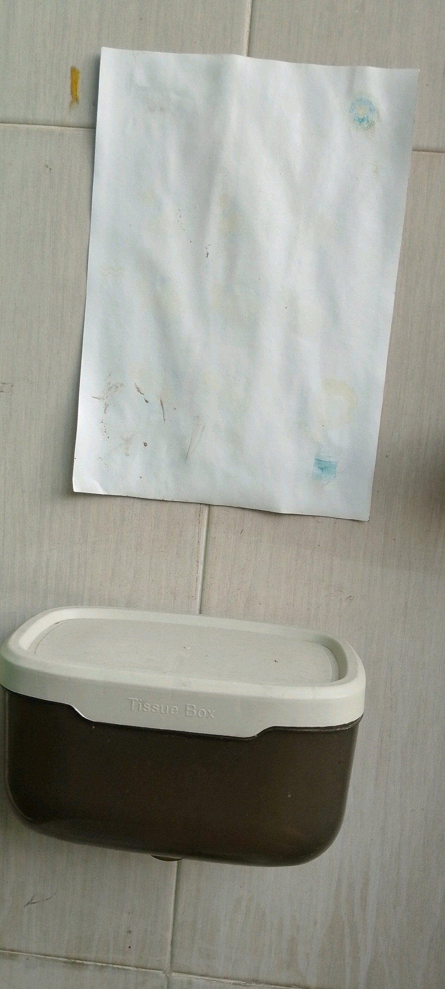 An overhead shot captures a tissue box with a beige lid and a dark brown base resting on a tiled surface, with a piece of crumpled white paper positioned above it. The paper appears to have some smudges and light blue markings, possibly from ink or paint. The tiled floor, a common sight in residential and commercial spaces in City of Medan, Indonesia, features light-colored square tiles with darker grout lines. The tissue box is oriented horizontally, and the words "Tissue Box" are subtly embossed on the beige lid. The scene is illuminated by natural light, casting subtle shadows. The composition suggests an indoor setting, perhaps a bathroom or a common area, where these items are used for everyday convenience.