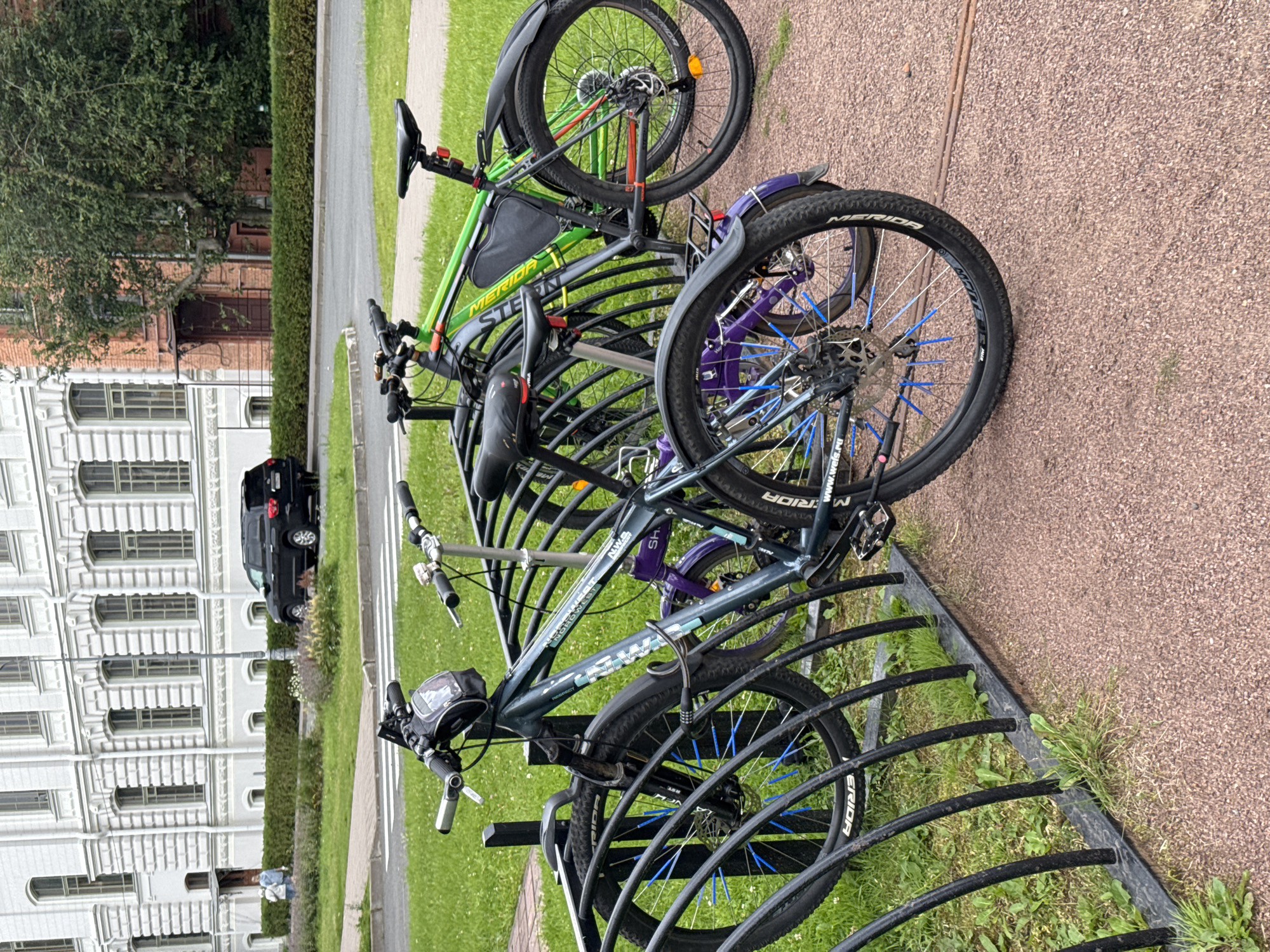 The image depicts an outdoor bicycle parking area adjacent to a building, likely in the city of Ladozhskoe Ozero, Russia. In the foreground, several bicycles are parked in a black metal rack. The bicycles vary in color, with one being dark gray, another green, and a smaller one purple. Notable details on the bicycles include colorful blue spokes on the wheels of the dark gray bike and a digital lock. The ground surface is a mix of red gravel and green grass. In the background, a black SUV is parked on a paved road, with a neatly trimmed hedge running alongside it. Behind the hedge, a large, light-colored building with numerous windows is visible. The overall scene suggests a public space within an urban setting.