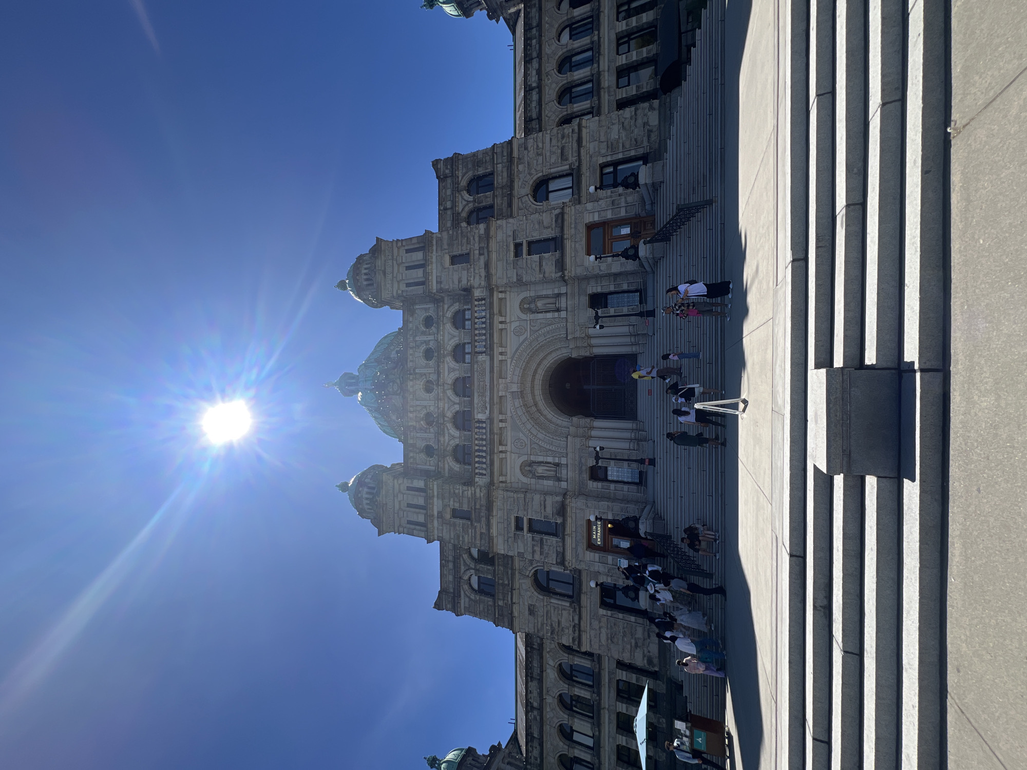 The image shows the exterior of the British Columbia Parliament Buildings, a large stone building with domes, located in Victoria, Canada. The sun is high in the sky, creating a bright glare that partially obscures the top of the image. A wide set of stone steps leads up to the main entrance of the building, where several people are seen walking and standing. Some individuals are gathered on the steps, while others are further up, approaching the entrance. The building exhibits classical architecture with arched windows and decorative stonework. To the left of the steps, there is a light blue umbrella and a cart. The foreground consists of more stone steps and a paved area. The scene is bathed in bright daylight, suggesting a clear and sunny day.