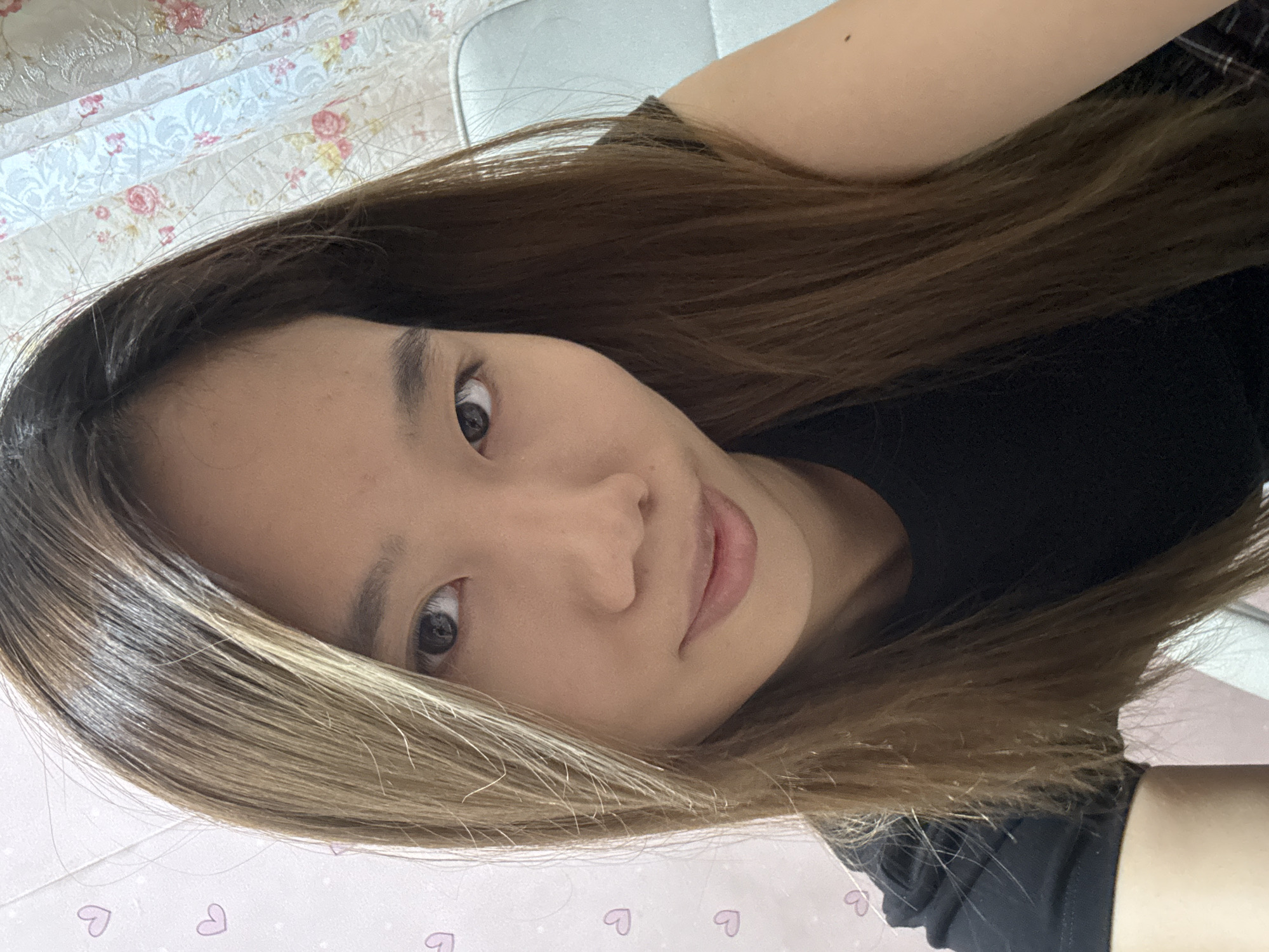 A young woman with long, straight brown hair with blonde highlights is shown from the chest up, taking a selfie. She is wearing a black t-shirt. Her left eye is visible, and she has a slight smile. The background consists of a pink wall with faint heart decorations and a curtain with a floral pattern. The scene appears to be indoors.