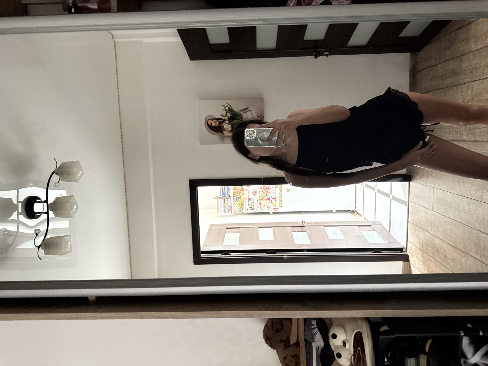 A young woman, with dark hair braided to her left side, is taking a selfie in a mirror. She is wearing a black tank top and black lace-trimmed shorts. She is holding a silver smartphone in her right hand, obscuring her face. The background of the reflection shows an open door revealing a hallway with a checkered floor and another door further down. To the left of the door, there is a framed picture of a woman wearing a hat and holding tulips. The room appears to be a bedroom or dressing area, with a tall mirror on the right, a closet door on the far right, and a shelf with stuffed animals and clothing on the far left. The flooring is light-colored wood. The lighting is bright, suggesting daytime. The overall scene is casual and personal, indicative of a private residence.