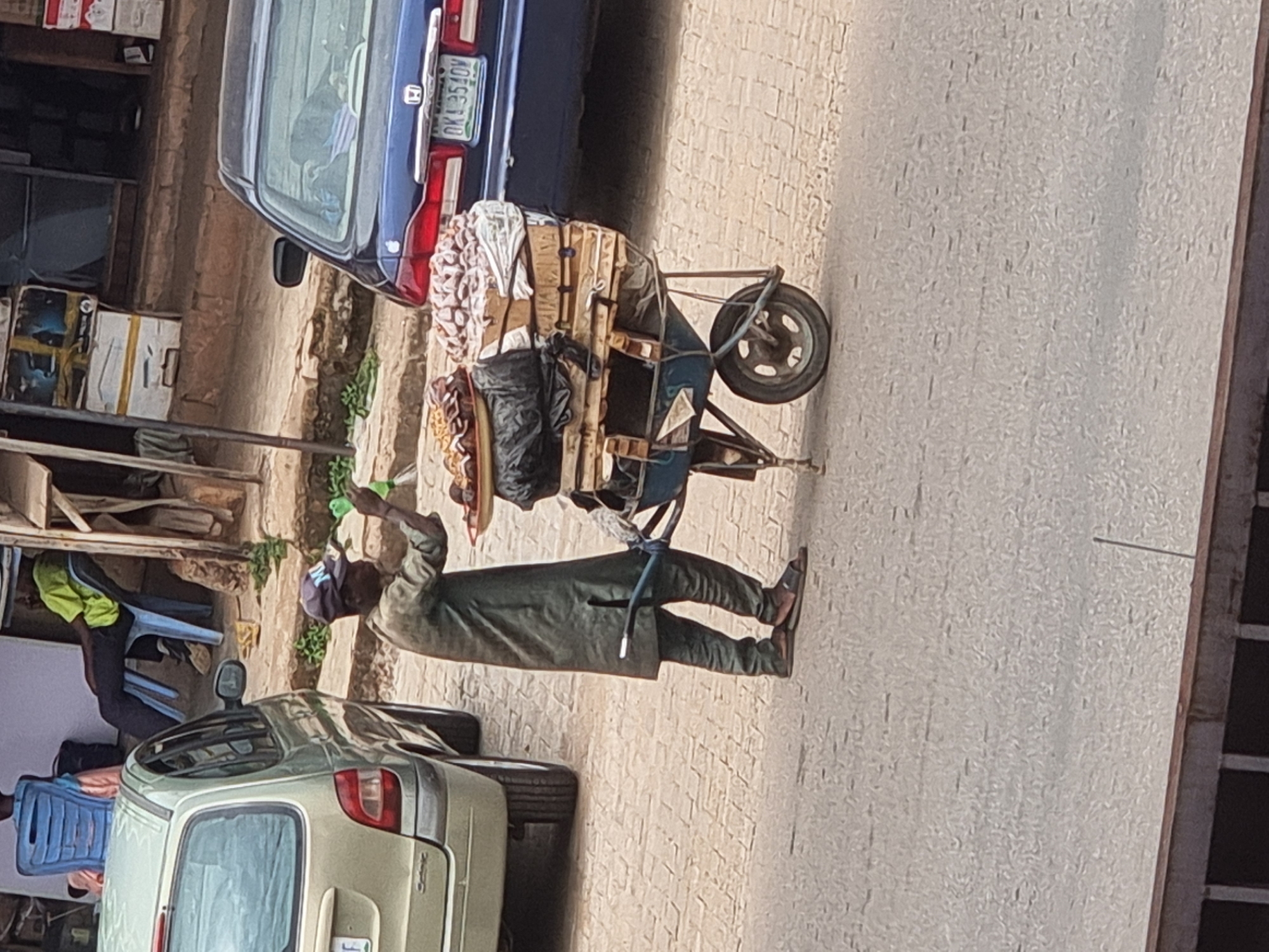 A man pulls a heavily loaded wheelbarrow along a paved street in Jos, Nigeria. The wheelbarrow contains sacks and items wrapped in white cloth, stacked on top of a wooden frame. A blue car is parked nearby with the license plate "OKA 3510V". Another car is parked behind the man. There are buildings on either side of the street and individuals are seen in plastic chairs near a shop. The man is wearing a grey cap, a dark shirt and pants, and sandals. He is holding a green plastic bottle.