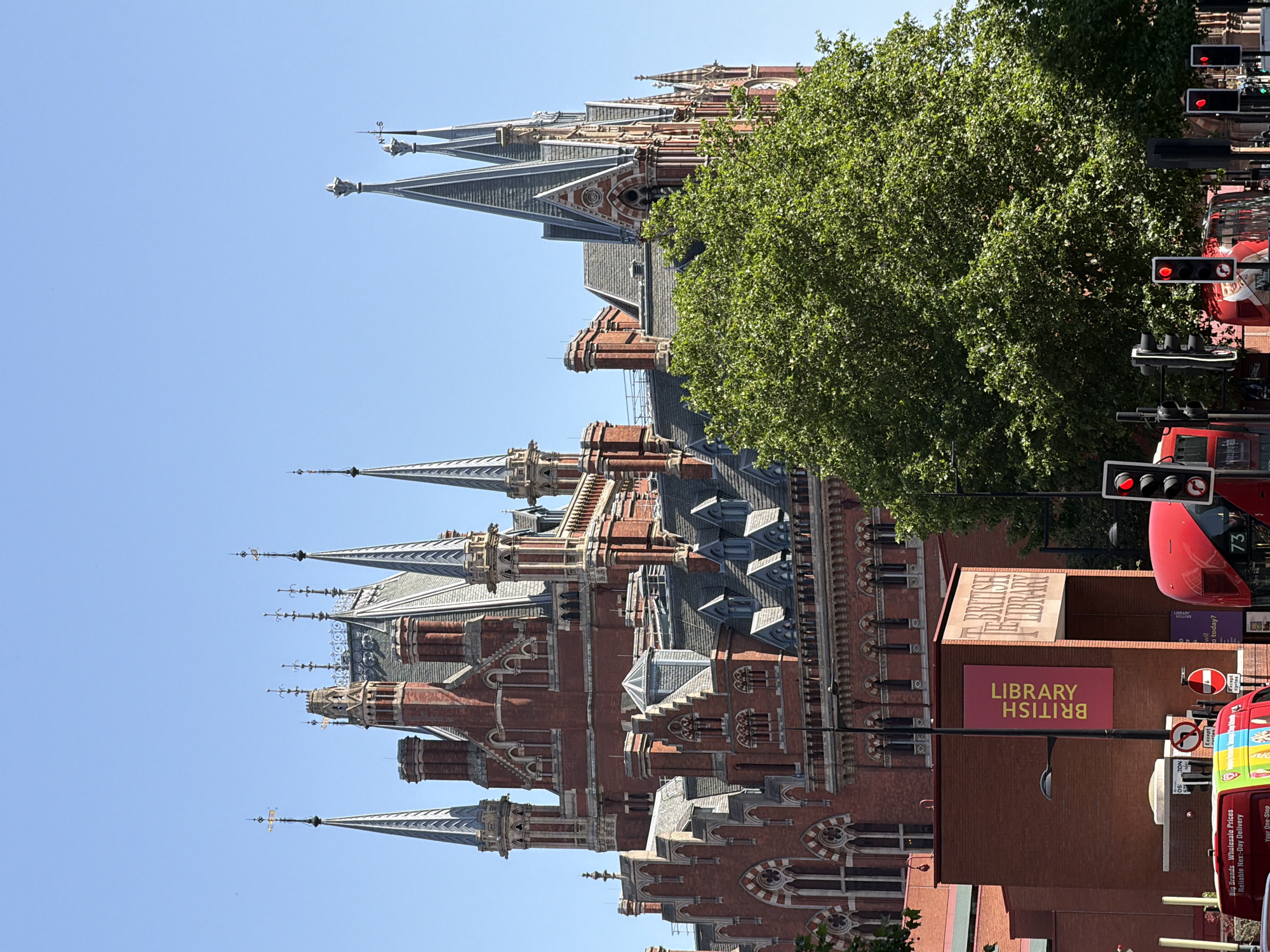 The image shows St Pancras railway station in London, United Kingdom, with a red bus and a sign for the British Library in the foreground. The station is a large, ornate Victorian building with multiple spires. The sky is blue and clear. A large tree is visible in the middle ground, partially obscuring the station. Traffic lights show red, indicating a stop. Several red double-decker buses are present on the road. The British Library signage is prominent.
