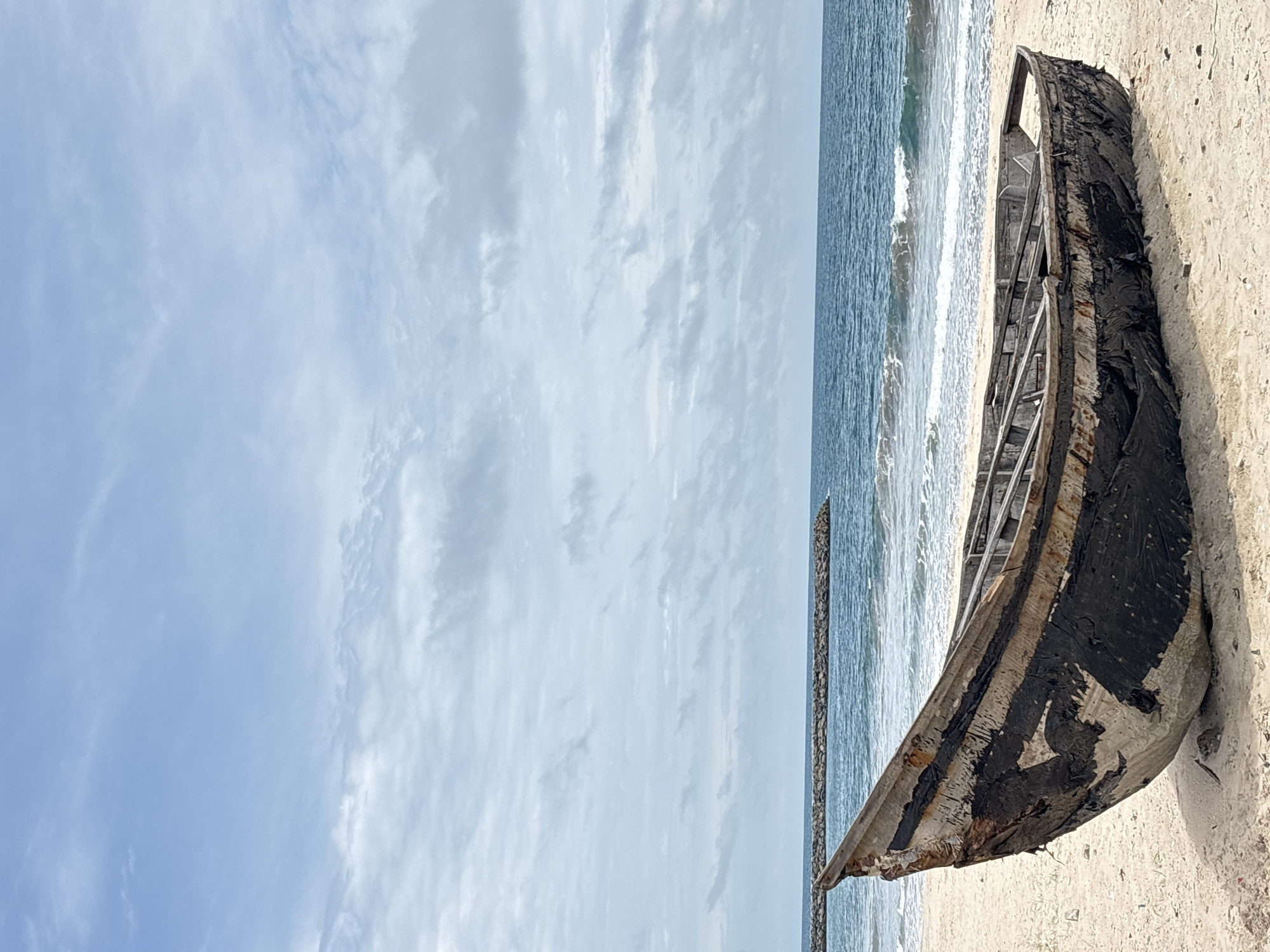 The image captures a weathered, abandoned wooden boat lying on a sandy beach in Alaguntan, Nigeria. The boat is predominantly dark in color with significant wear and tear, suggesting prolonged exposure to the elements. The beach is adjacent to the ocean, with waves visible breaking along the shoreline. In the background, a breakwater extends into the sea. The sky is overcast with scattered clouds. No people are visible in the scene. The overall atmosphere is one of solitude and decay.
