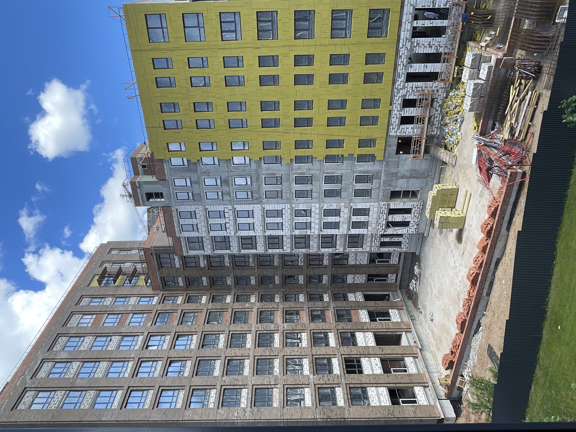 A construction site in an urban environment features several multi-story buildings under various stages of construction. The building on the left is clad in dark brown brick and has numerous windows covered with plastic sheeting. Adjacent to it, another building is under construction with exposed grey blocks and scaffolding. To the right, a building is in a more advanced state, with yellow insulation panels covering its exterior, and many windows are visible. The ground level of the construction site is a mix of sand, concrete, and piles of building materials, including bricks, rebar, and wooden planks. A dark metal fence encloses part of the site, with a small patch of green grass visible beyond it. The sky above is blue with scattered white clouds.