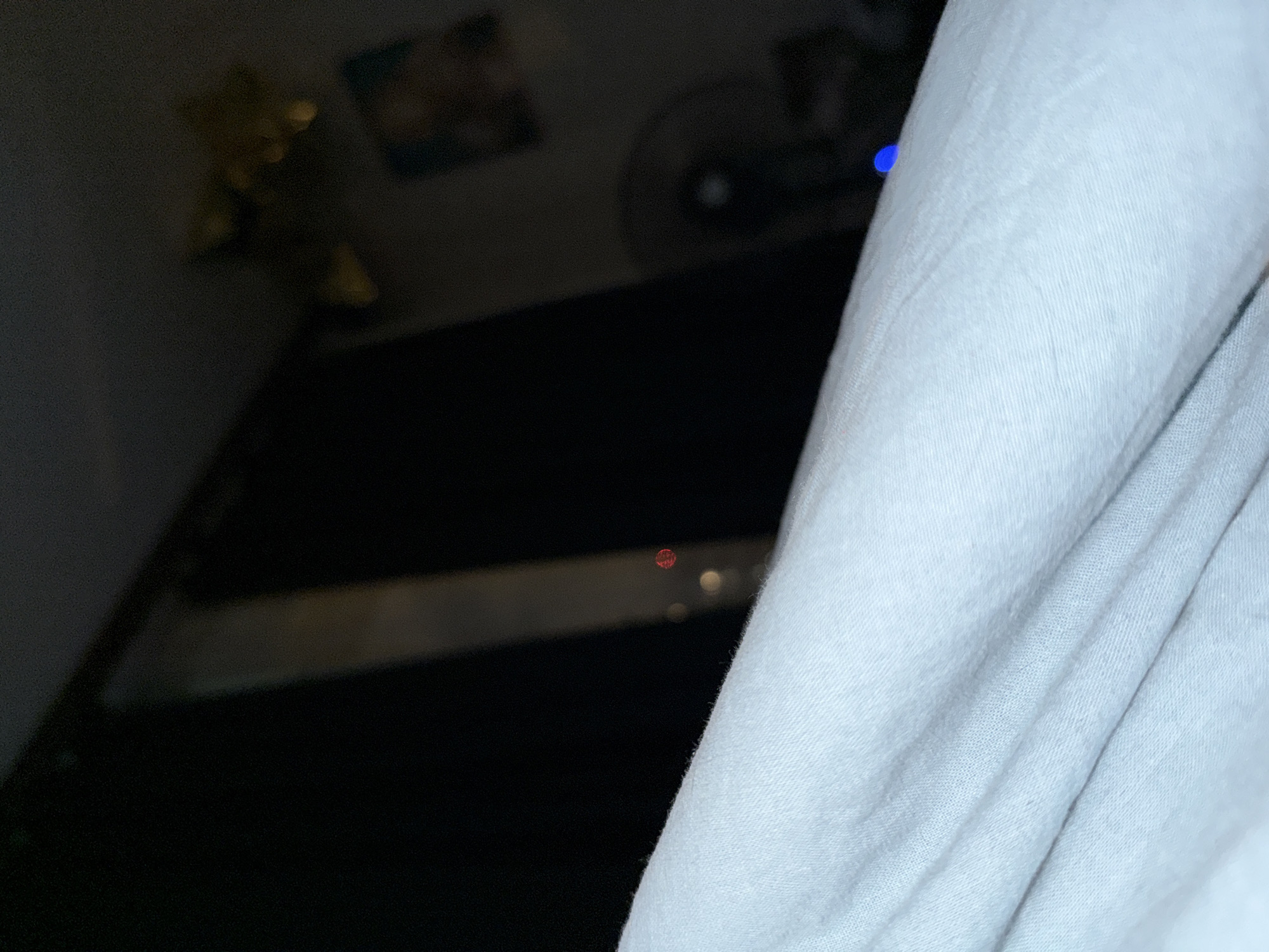 The image shows a white fabric, likely a bedsheet or blanket, dominating the right side of the frame. The background is dark and out of focus, but suggests an interior setting. A red LED is faintly visible in the midground, possibly from a device. Other indistinct shapes and lights appear in the background. The location (Khimki, Russia) cannot be determined from the visual content alone. There are no people visible.