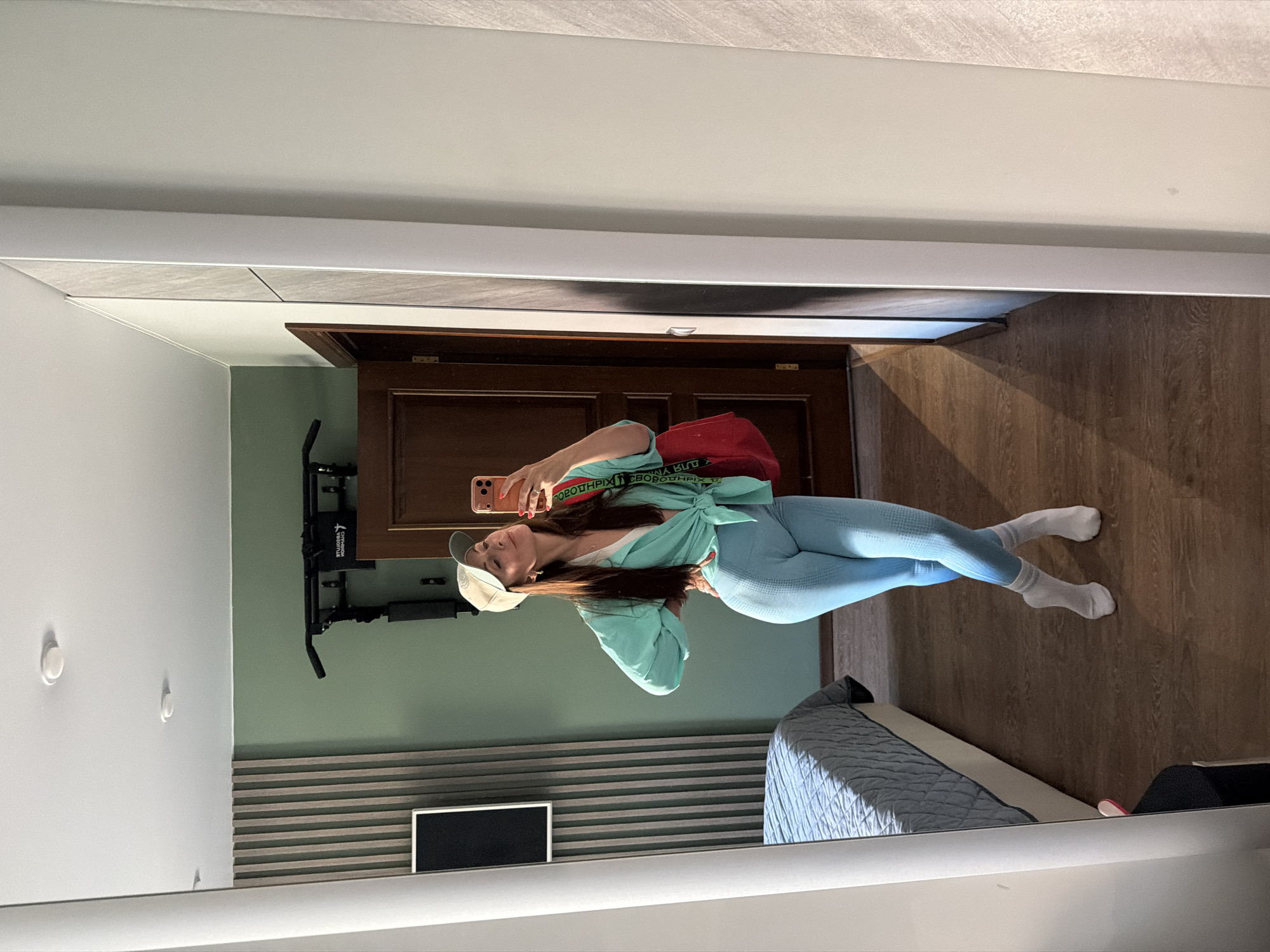 A woman stands in a bedroom, taking a mirror selfie. She is wearing a light blue long-sleeved shirt tied at the waist, light blue leggings, white socks, and a beige baseball cap. A red bag with black straps is slung over her shoulder. The room has a bed with a blue and grey quilted comforter, a wooden door, a pull-up bar mounted on the wall, and a television screen. The walls are painted light green and textured beige, and the floor is dark wood. The overall impression is of an informal, domestic setting. The image does not contain any information to suggest the location is Birobidzhan, Russia.