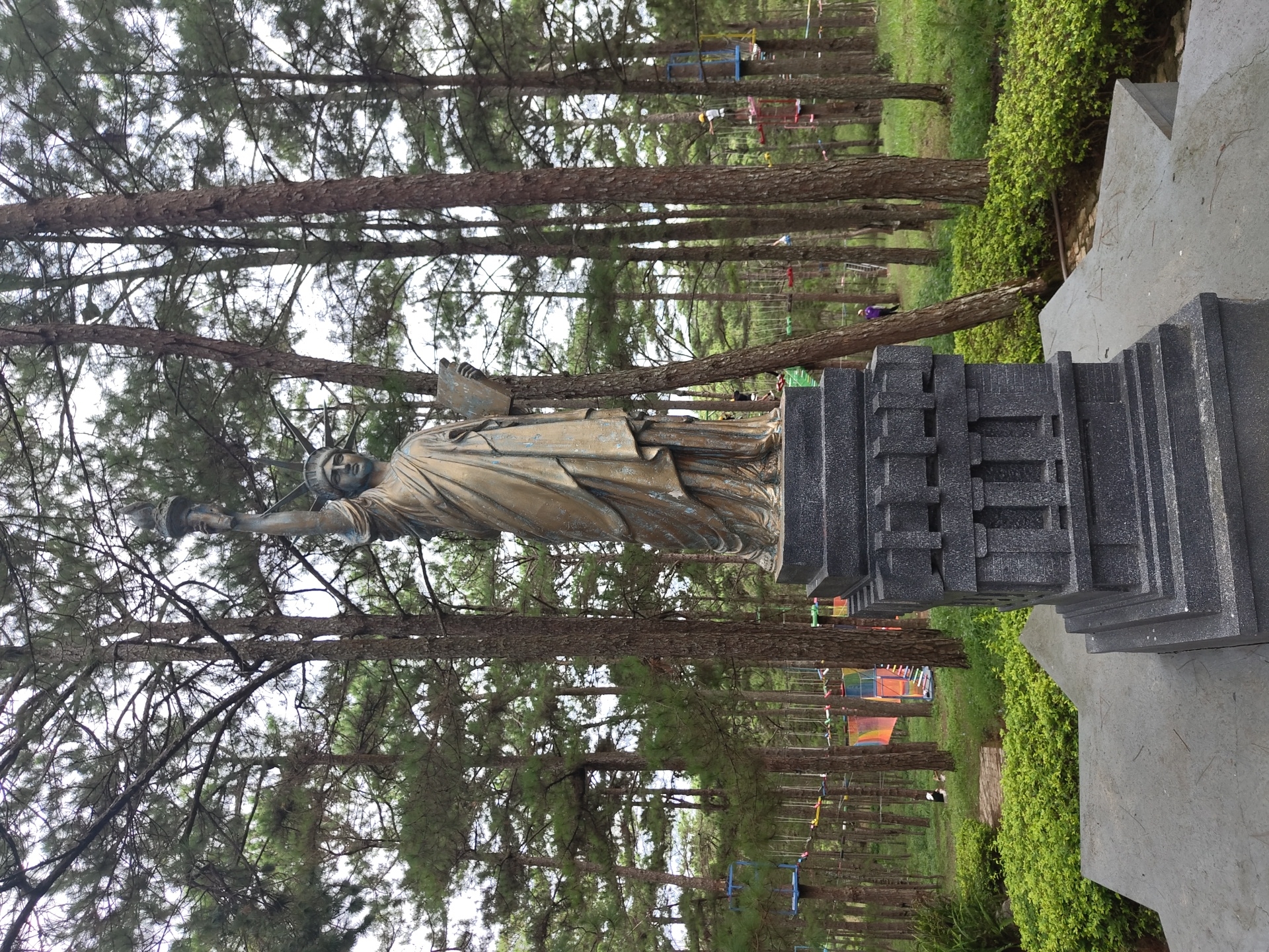 A bronze-colored statue of the Statue of Liberty stands on a dark gray pedestal in the foreground. The statue is approximately life-sized and faces forward. It holds a torch in its right hand and a book in its left. The pedestal is rectangular with carved architectural details. Behind the statue, a dense forest of pine trees stretches into the background. In the mid-ground, a rope course with colorful platforms and zip lines is visible, suggesting it's a recreational area. Several people are present in the background, some participating in the rope course, others walking. The ground is covered in green grass and lush bushes. The sky is overcast. The location appears to be Lâm Viên - Đà Lạt, Vietnam, known for its pine forests and adventure parks.