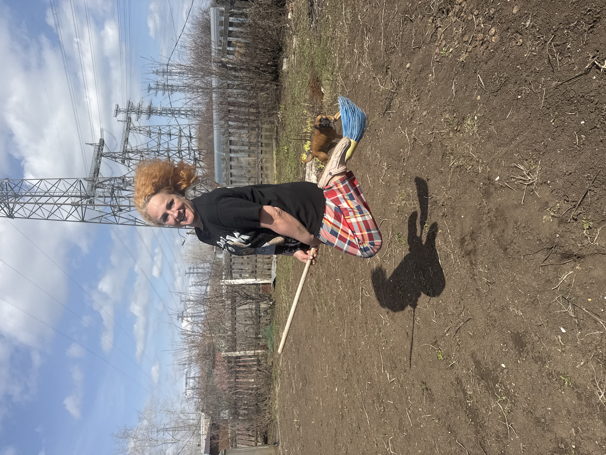 A woman with reddish-orange hair is in a yard in Chaikovsky, Russia, holding a wooden stick. She wears a black shirt, a plaid skirt, and white Crocs with a blue broom attached to one. A dog stands nearby. Power lines are visible in the background, along with bare trees and a fence. The ground is dirt.