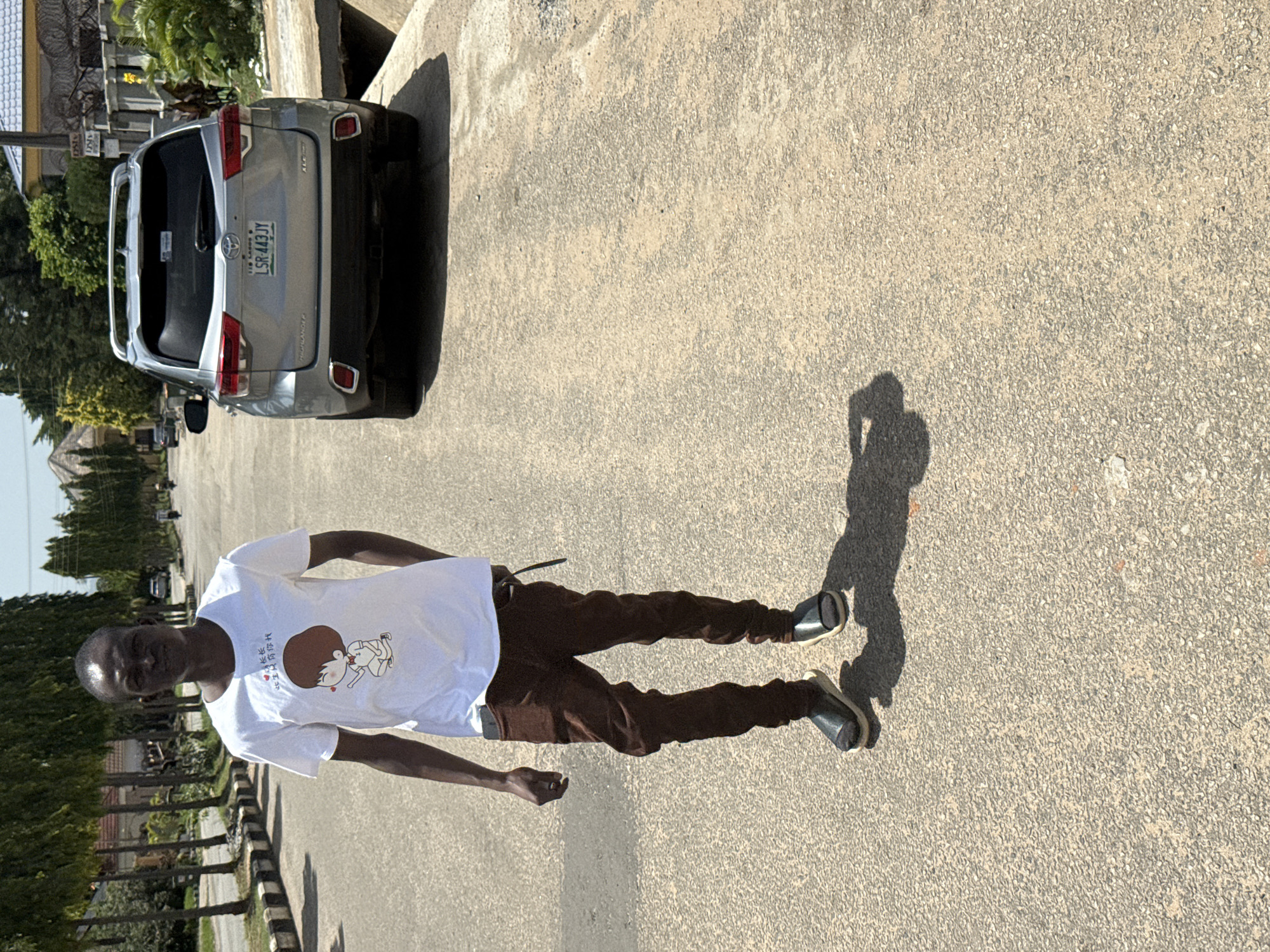 A man in a white t-shirt and brown pants stands on an asphalt road near a silver SUV with license plate LSR-443JY. The location is in Abuja, Nigeria, as indicated on the license plate. Trees line the side of the road. The man's shadow is cast on the road.