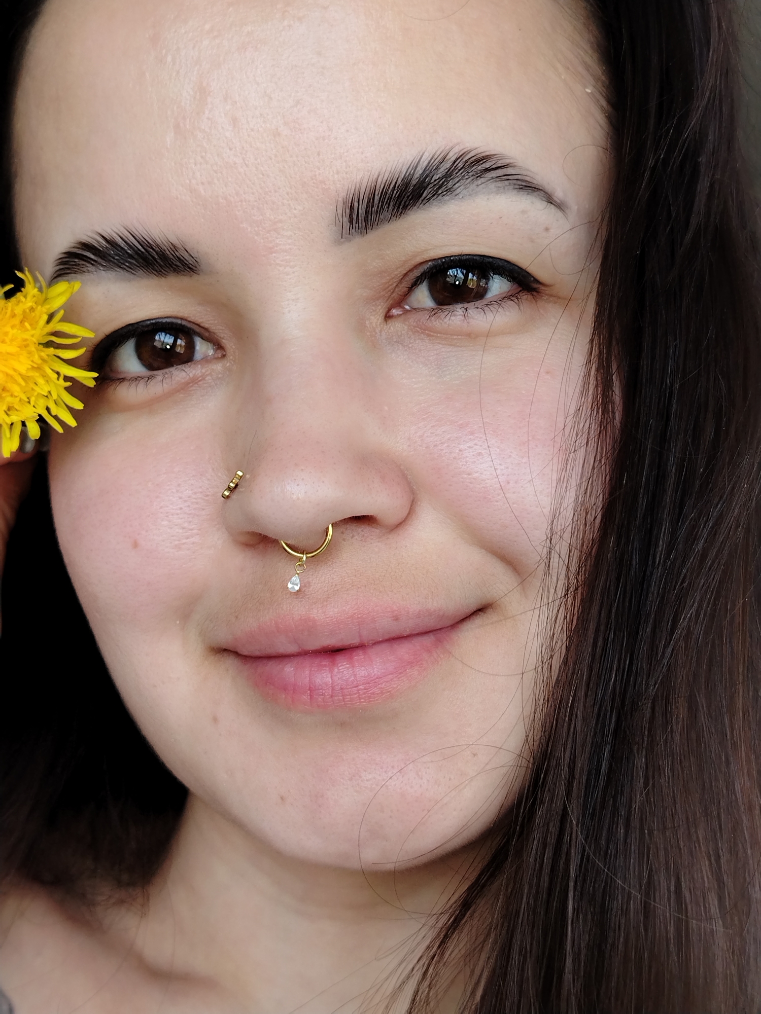 A close-up, portrait-style photograph shows a person with light skin and dark brown hair, likely female. The person has brown eyes and well-defined eyebrows. The right side of their face is more visible, revealing a partial smile. Two pieces of gold nose jewelry are visible: one is a stud on the left side of the nose and the other is a ring with a dangling teardrop-shaped gem in the center. A yellow dandelion is partially visible to the left of the face, near the eye. The background is dark and indistinct. The location of Noviy, Russia is not confirmed by the image.