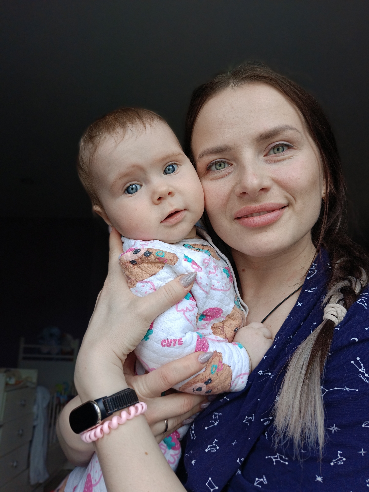 A fair-skinned woman with green eyes and brown hair styled in a braid is holding a fair-skinned baby with blue eyes. The baby is wearing a white outfit with cartoon animals printed on it, and the woman is wearing a dark blue garment with a star pattern. The woman has a black fitness tracker and pink hair tie on her left wrist. The background is partially blurred and consists of a dark wall and some furniture, suggesting an indoor setting. There is no information available to indicate the location is in the city of Rusalevka, Russia.