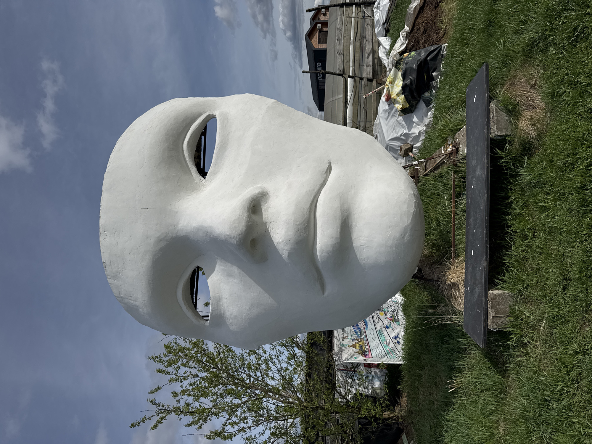 A large, white, hollow face sculpture is the focal point of the image, positioned in a grassy area under a partly cloudy sky. The sculpture has eye holes, a nose, and a mouth, creating a human-like visage. Behind the face, construction or building materials are visible, including wood structures, a dark container with the words "TAC ПИЛО" visible on it, and what appears to be covering material. To the left of the face, there is a small tree. A black rectangular sign is positioned to the right of the mask. In the background, there is a structure with dark roof and siding and wooden fencing. The scene suggests an outdoor installation in Nizhny Novgorod, Russia.