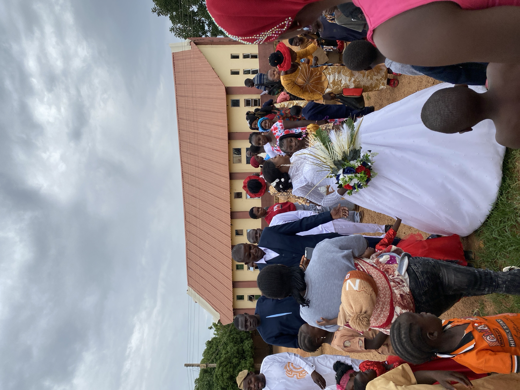A daytime outdoor shot depicts a wedding gathering in Kuru Dungus, Nigeria. A bride in a white gown stands amongst a crowd of people dressed in a variety of attire, from formal suits to traditional African clothing. The setting is a dirt surface next to a building with a red roof. The sky is overcast with grey clouds.
