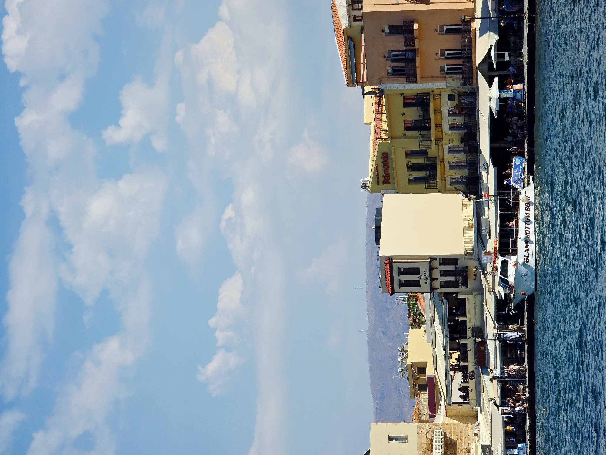 This is a photograph of the harbor in Chania, Greece, taken on a clear, sunny day. The image showcases a waterfront scene with several buildings lining the harbor, most of which appear to be restaurants and shops with outdoor seating. The architecture is a mix of traditional and more modern styles, with colorful facades and tiled roofs.
In the foreground, the blue water of the harbor is visible, with gentle waves. A white boat, labeled "GLASS BOTTOM BOAT," is docked at the harbor's edge. Several people are on the boat, and more are seen on the boardwalk in front of the buildings. The people on the boardwalk are engaged in various activities, such as walking, sitting at tables, and conversing. Some are dressed in casual summer clothing.
Behind the buildings, a chain of hills or mountains forms the backdrop, with some visible telecommunication towers on the highest peaks. The sky above is a bright blue, dotted with fluffy white clouds. The lighting suggests it is daytime, likely late morning or early afternoon. The overall impression is a vibrant, bustling tourist destination.