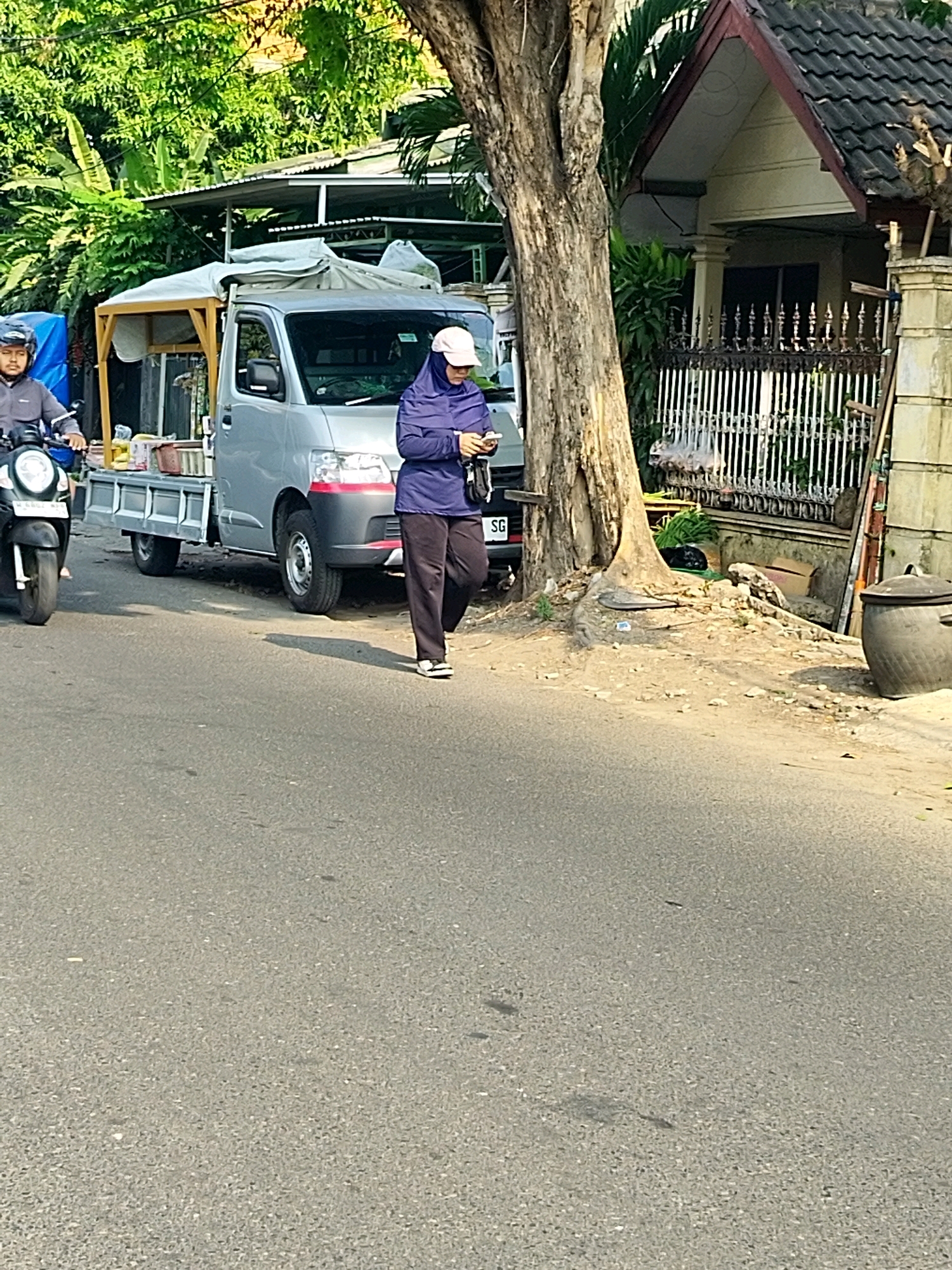 In Mojokerto, Indonesia, on a sunny day, a gray utility truck is parked on the side of a paved road. The truck has a canvas cover over its bed. A person wearing a purple jacket and a white headscarf is standing next to the truck, looking at their phone. To the left of the truck, a person on a motorcycle wearing a helmet is also visible. The surrounding environment includes a large tree with exposed roots, lush green foliage, and a residential building with a white fence. The road surface appears to be asphalt.