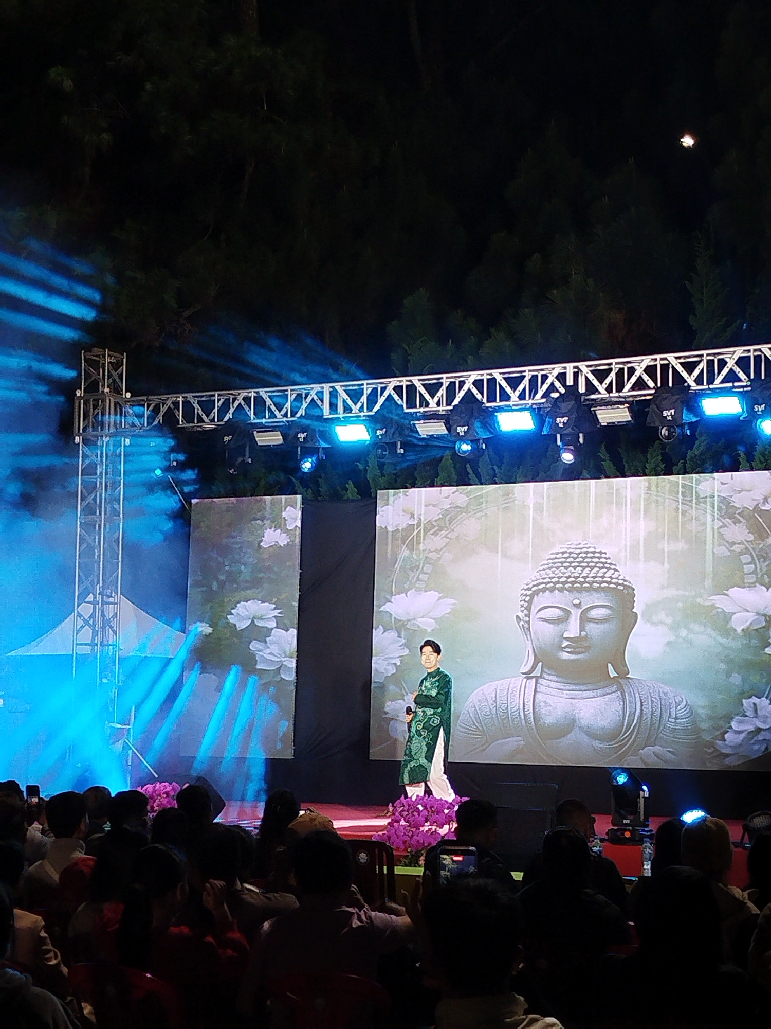 The image shows a scene in Da Lat, Vietnam, featuring a stage with lighting and a screen. A person stands on the stage, with a backdrop of a Buddha statue on the screen. An audience is present. A metal structure with lighting is positioned above the stage. The environment is dark, potentially at night, with trees visible in the background.