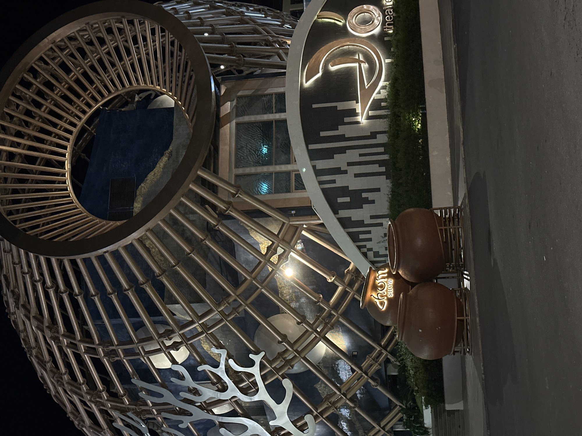 This media file showcases an exterior view at night, likely in Nha Trang, Vietnam. The scene features a modern building with a distinctive architectural design. A prominent element is a large, spherical structure made of interconnected metallic rods, with a circular opening revealing a dark, textured surface behind it. Adjacent to this is a dark, curved facade with a brightly lit, stylized logo featuring a circular element and a curved line, accompanied by the word "theatre" in white text. Below this is a patterned wall with horizontal lines in alternating black and white. In the foreground, three large, brown, pot-like objects are stacked vertically, each illuminated with a glowing "chom SHOW" sign. Lush green bushes are visible behind these pots and along the base of the patterned wall, suggesting an outdoor setting. The ground appears to be asphalt, possibly a sidewalk or road, with some reflections indicating a damp surface, suggesting recent rain or a humid environment. The lighting is artificial, with the building's signage and spotlights creating a dramatic effect against the dark night sky. There are no people visible in the frame, and the overall mood is one of modern aesthetics and evening ambiance. The prominent "theatre" text on the building facade suggests a venue for performances.