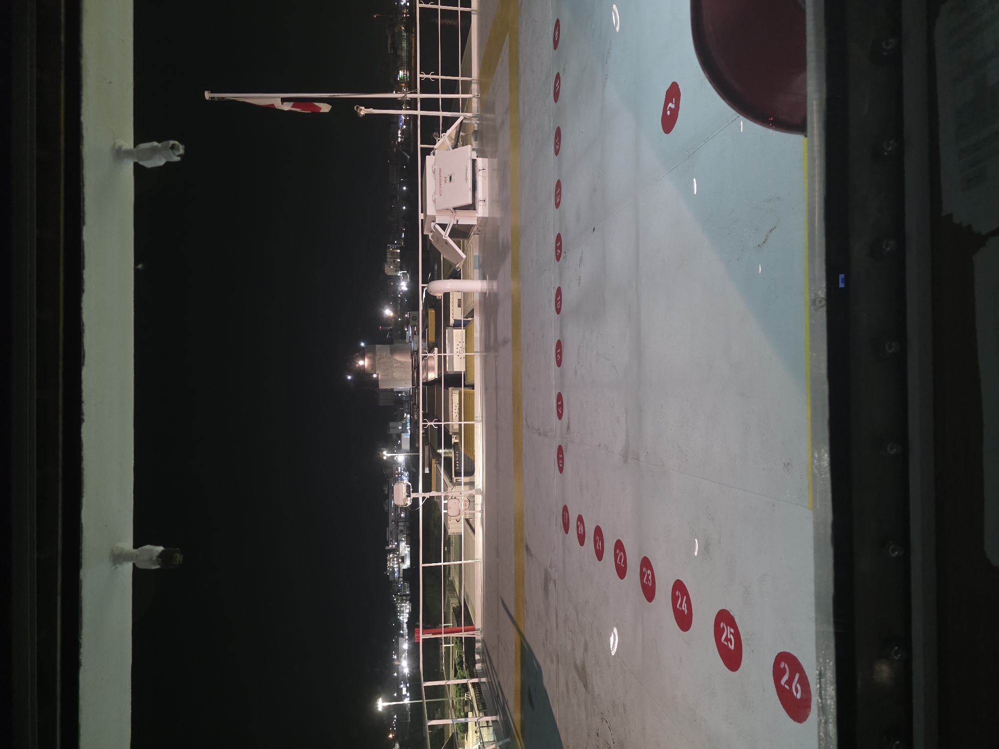 The image shows a deck at night, likely on a ferry, with numbered red circles painted on the deck. The circles are numbered sequentially, starting from 26 and going up to 16 on the left side of the image, and from 2 to 1 on the right side. In the background, a city skyline is visible with lights from buildings and streetlights. There is a flag pole with a flag on the right side of the image. A red chair is partially visible in the bottom right corner. The scene is well-lit by artificial lights. The overall environment suggests a transportation hub or a maritime setting. The numbered circles are likely used for vehicle or passenger placement. The city in the background is Brindisi, Italy, as indicated by the port area and city lights characteristic of Brindisi at night.