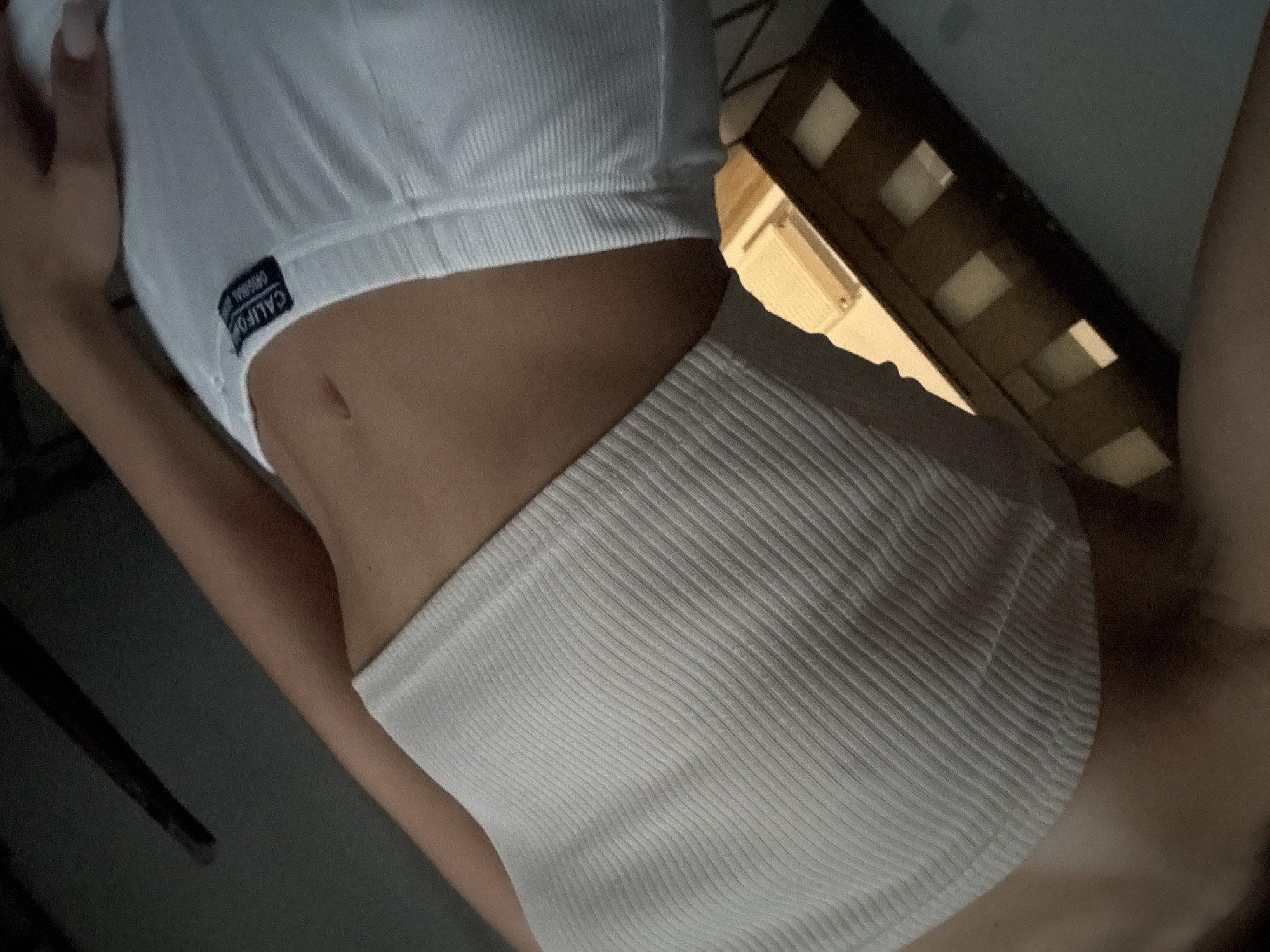 The image is a close-up, low-angle shot of a person's midriff. They are wearing a white ribbed crop top and white shorts with a label that reads "CALIFORNIA ORIGINAL". The background is dark and out of focus, with a wooden structure visible at the bottom right, possibly part of furniture or a window frame. The lighting is dim, casting shadows. The setting appears to be indoors, and there is no specific indication of a location in Chișinău, Moldova.