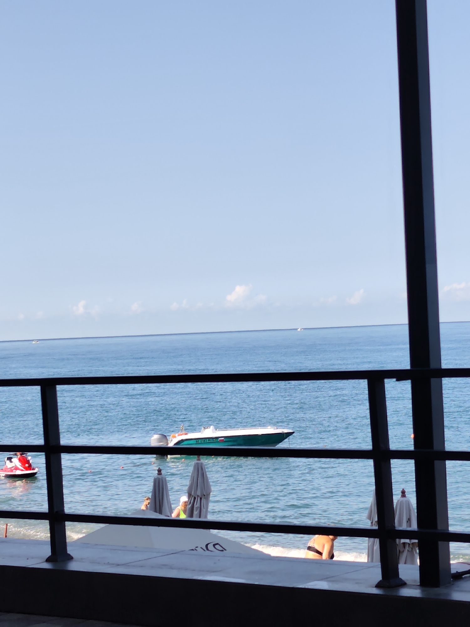 This image captures a tranquil seaside scene with the Black Sea stretching out under a clear blue sky. In the foreground, a dark metal railing frames the view, suggesting an elevated vantage point, possibly a balcony or a shaded seating area. Beyond the railing, the gentle waves of the sea are visible, with a small white boat anchored in the distance. Closer to the shore, a red jet ski is partially submerged in the water, and a white speed boat with a green hull is moored. Two folded beach umbrellas stand on the sandy shore, with several beachgoers enjoying the water and the sun. The overall ambiance is one of a relaxed summer day at the beach in Sochi, Russia, a popular resort city known for its picturesque coastline.