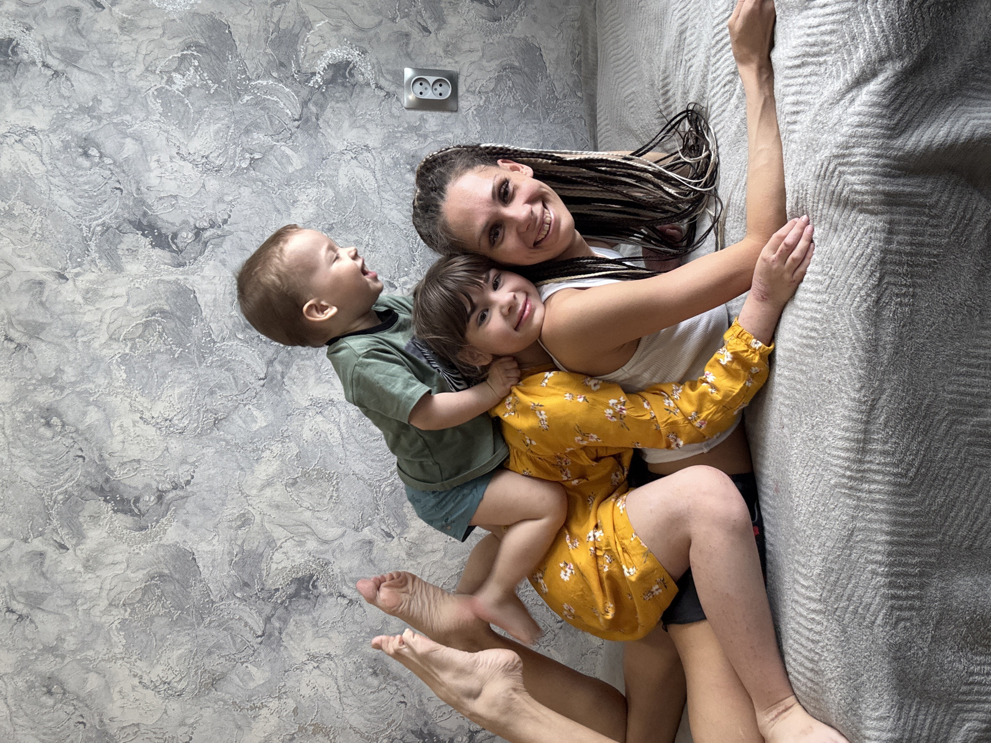 The image captures a joyful family moment featuring a woman and two children playfully posing indoors, likely in Rusanovo, Russia. The overhead shot shows them lying on a textured grey surface, resembling a bed or sofa, positioned alongside a decorative grey wall.
The woman, appearing to be the mother, lies on her back with her head turned to the left, smiling brightly at the camera. She has long, dark braided hair with lighter highlights and wears a white tank top. An older child, a girl, is nestled close to the woman, resting her head on the woman's shoulder and also smiling at the camera. She is dressed in a yellow long-sleeved dress with a subtle floral pattern.
A younger child, a boy, sits playfully on the older child's back, leaning towards the wall. His mouth is open wide, suggesting laughter or excitement as he looks upwards. He wears a green short-sleeved shirt and blue shorts.
The wall behind them features an intricate grey and white textured pattern, possibly decorative plaster. A silver electrical outlet with two sockets is visible on the wall near the woman's head. The grey surface they are lying on has a distinct chevron or zigzag pattern, characteristic of a cozy blanket or duvet. The scene conveys a warm, affectionate, and happy family interaction during what appears to be daytime. No text is visible in the image.