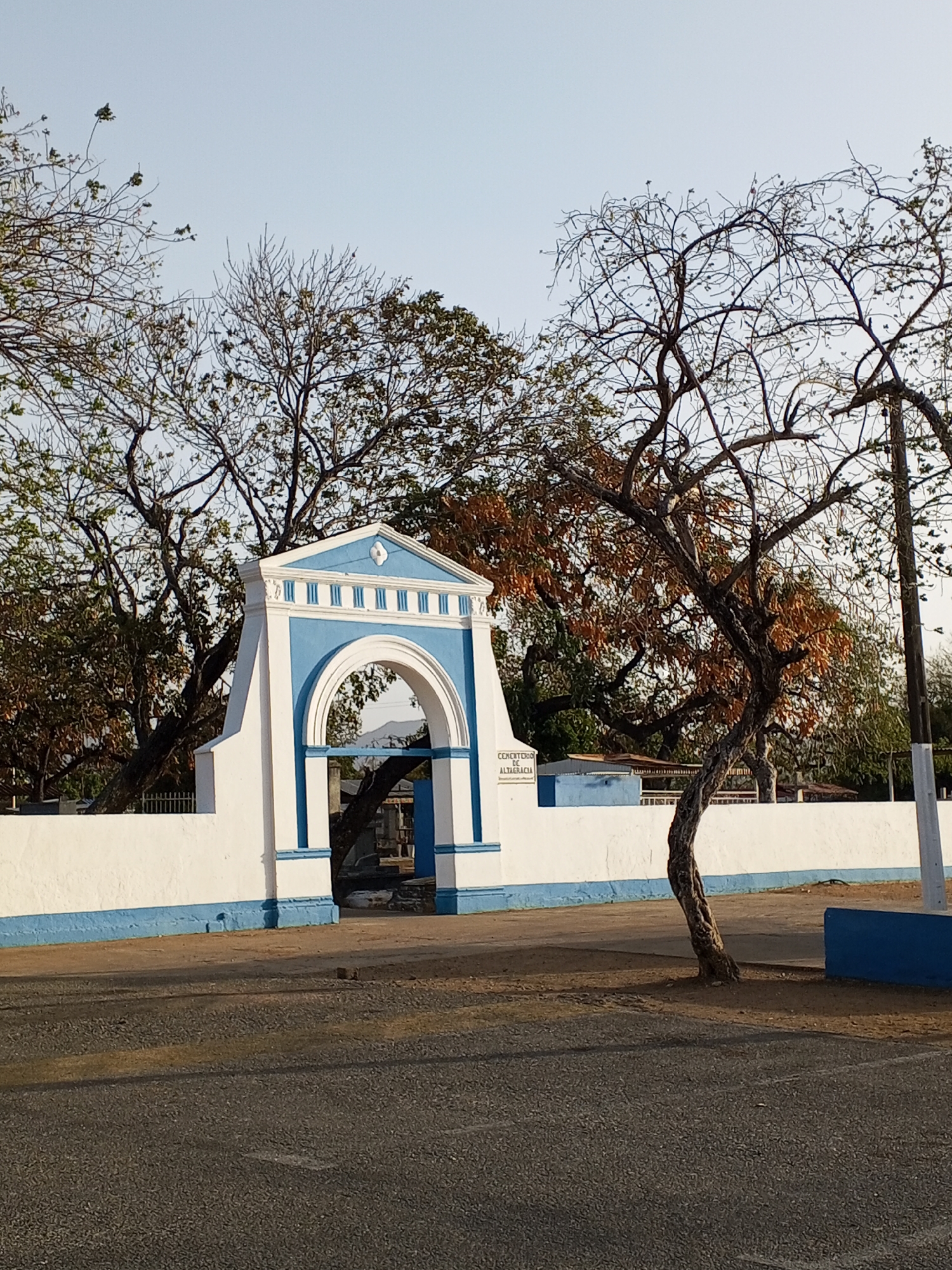 The image depicts the entrance to the Cementerio de Altagracia, a cemetery located in the city of Altagracia, Nicaragua. The entrance consists of a white wall with a blue stripe at the bottom and a white archway with blue trim. The archway is flanked by trees, some of which are bare. In the foreground is a paved road.