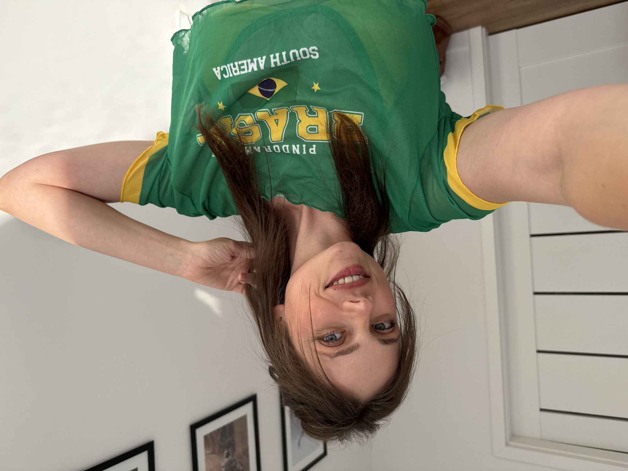A young woman with light brown hair and blue eyes is taking a selfie in an indoor setting. She is wearing a sheer green mesh top with yellow trim, which features the word "BRASIL" and the Brazilian flag printed on it, along with the text "SOUTH AMERICA". Her right arm is raised and her hand is touching her hair. The background includes a white wall, a white door with horizontal panels, and two framed pictures on the wall. The lighting suggests it is daytime, with sunlight casting shadows on the wall. The scene does not appear to be located in the city of Szklarska Poręba, Poland.
