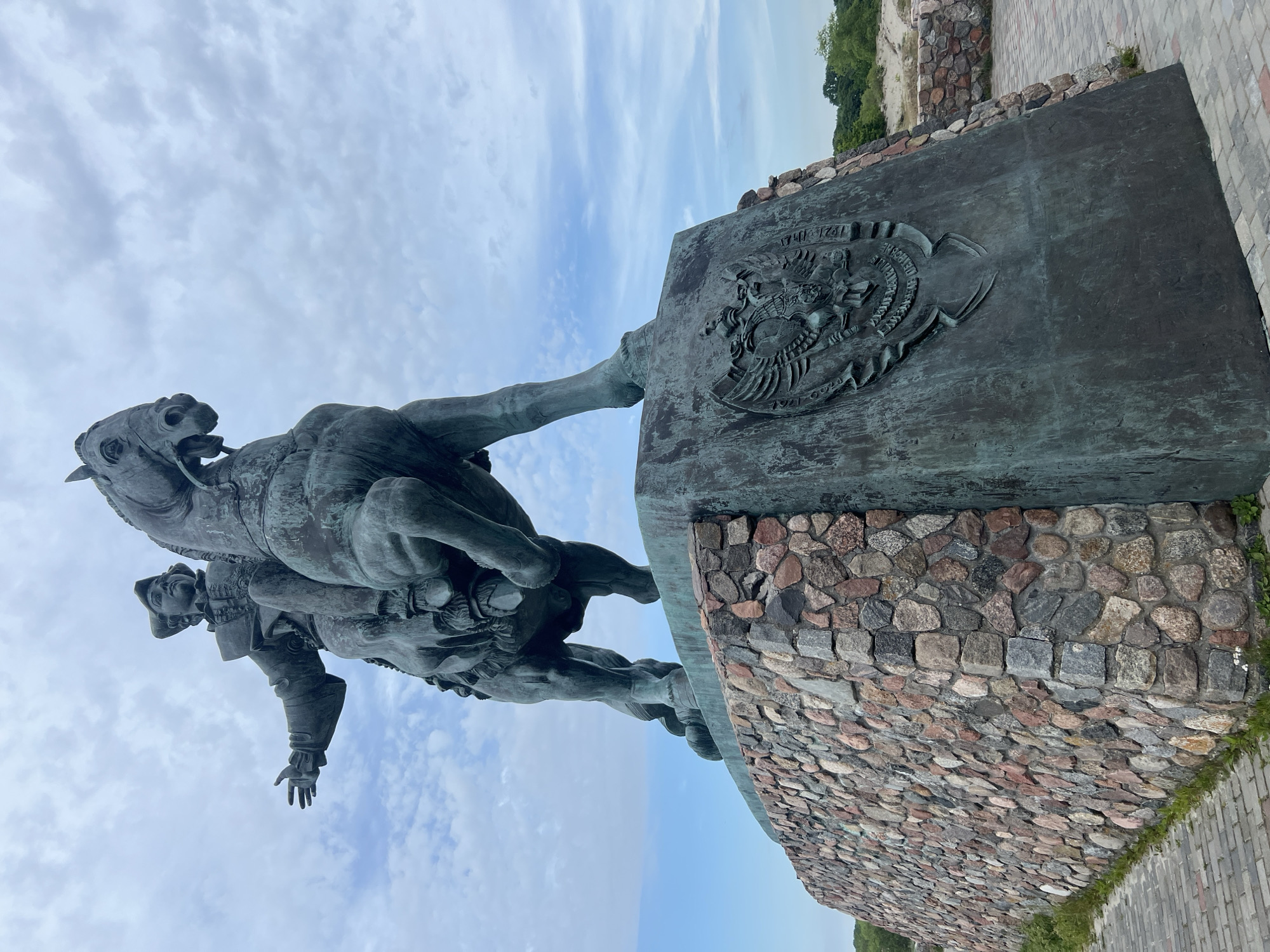 An equestrian statue of Peter I, also known as Peter the Great, stands on a stone pedestal. The bronze statue depicts Peter I in 18th-century attire, including a hat, a jacket with a ruffled collar, and breeches, riding a horse. He is gesturing with his left arm outstretched. The horse is depicted in mid-stride with its mouth open. The pedestal is constructed of rough-hewn stones of various colors, including gray, pink, and brown, forming a rough, uneven surface. A bronze plaque with a two-headed eagle emblem, possibly a coat of arms, is affixed to the front of the pedestal. The statue is located in Baltiysk, Russia, at the westernmost point of Kaliningrad Oblast. The background consists of a cloudy sky with patches of blue, and further in the distance, some sparse vegetation and sand dunes are visible, suggesting a coastal or open landscape.