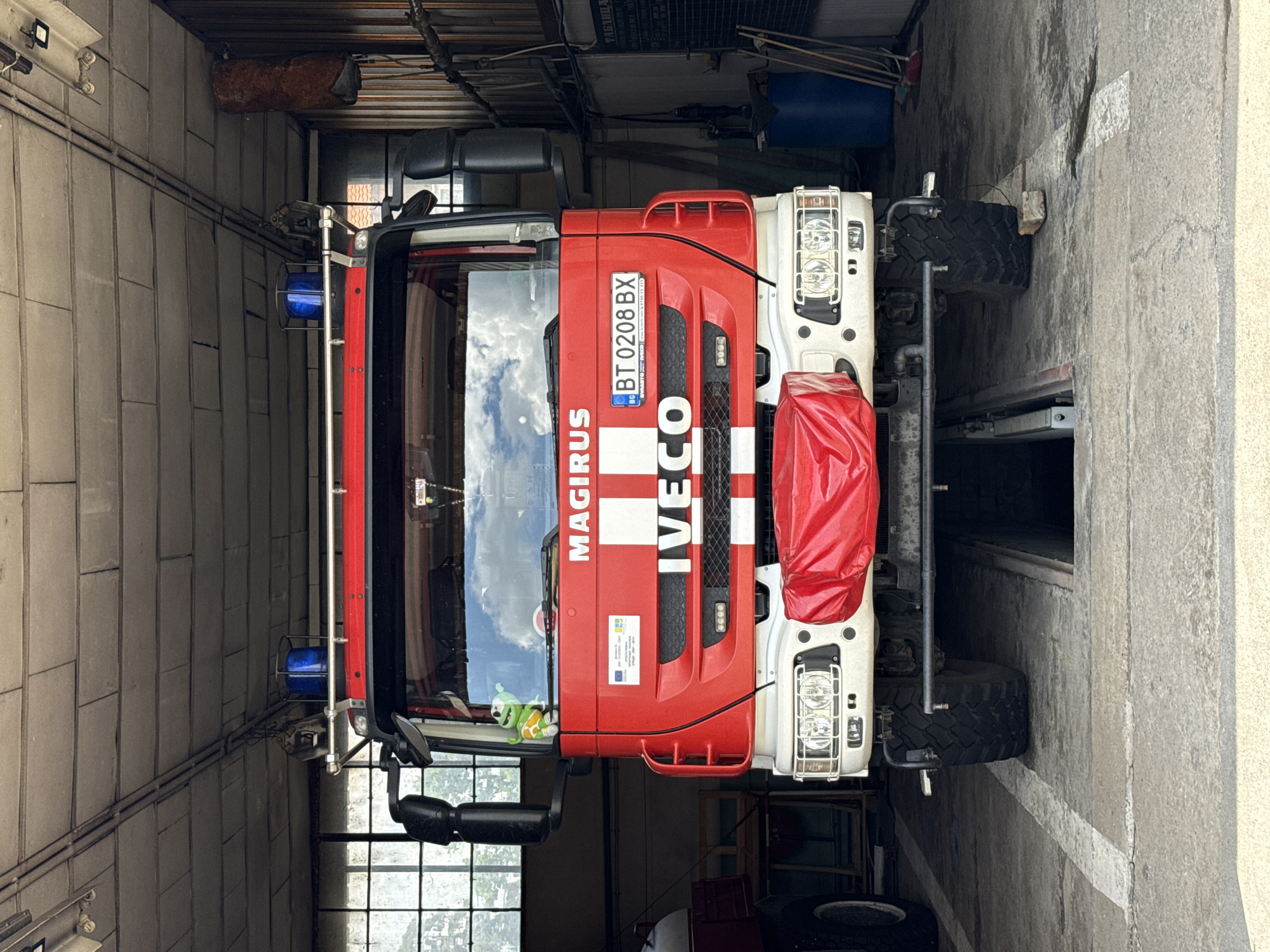 The image shows a red and white Iveco Magirus fire engine with Bulgarian license plate BT 0208 BX parked inside a garage with gray concrete walls. The location is likely Strazhitsa, Bulgaria, based on the license plate. The fire engine has emergency lights on its roof. The environment is somewhat cluttered. There are no people visible in the image.