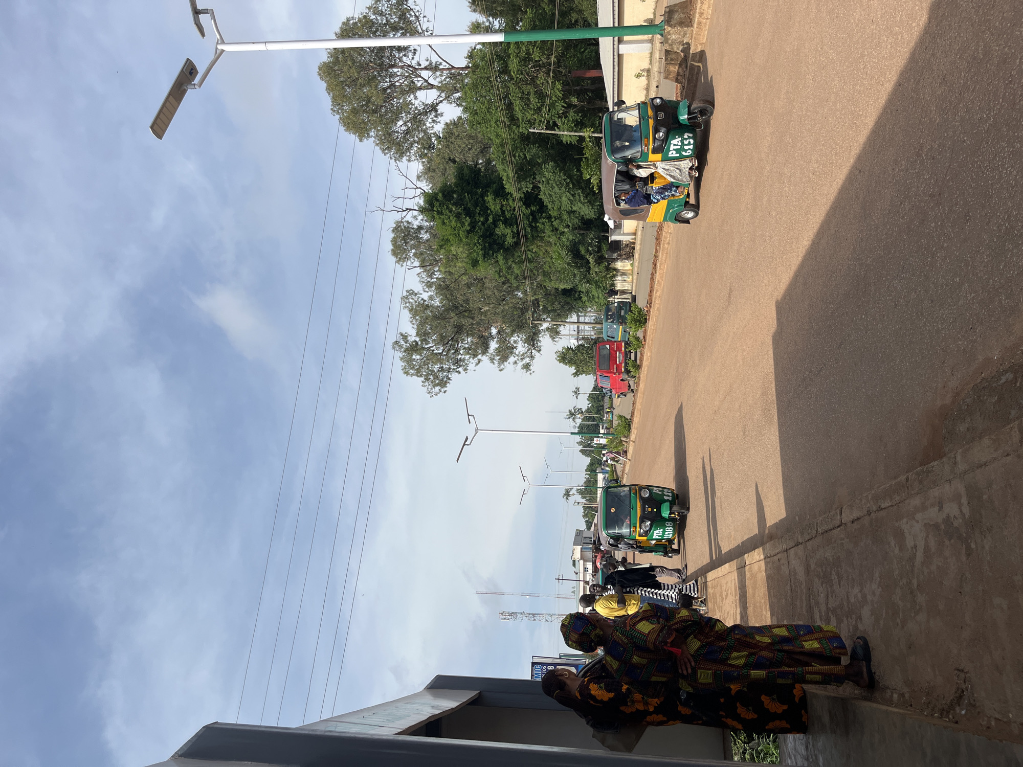 In Bukuru, Nigeria, this image captures a street scene under a partly cloudy sky. A group of people, including women in vibrantly patterned clothing and a man in a yellow shirt, stand on a raised concrete walkway along the side of a paved road. Their attention is directed towards the street. Two green and yellow auto-rickshaws, a common mode of transport in the region, are visible. One auto-rickshaw is stationary near the people on the walkway, while another is in motion further down the road. Trees line the street in the background, and a modern street light with solar panels stands tall. The road appears to be a main thoroughfare, with other vehicles and buildings in the distance. The overall impression is a snapshot of daily life in a Nigerian city.