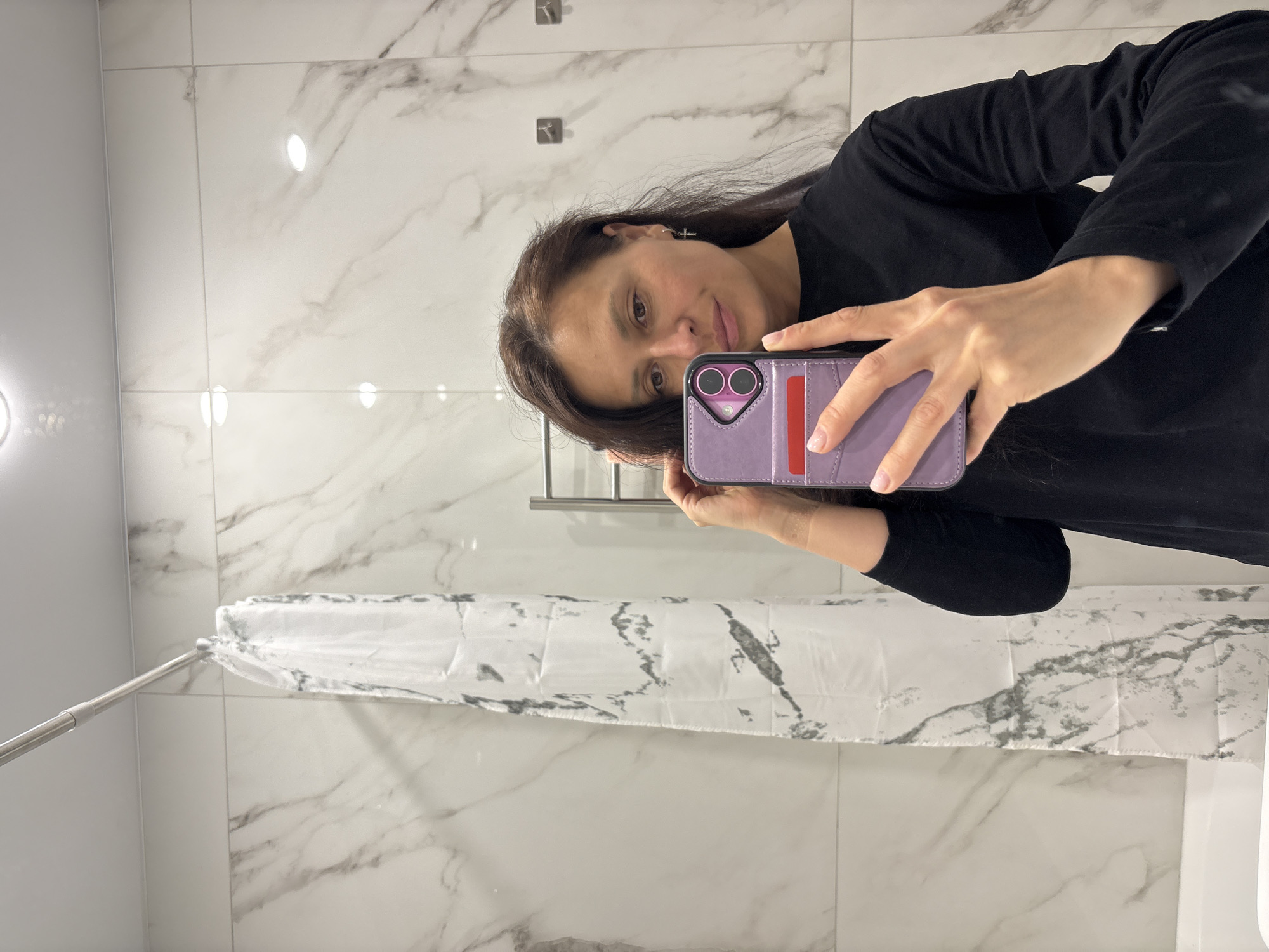 A woman is captured in a mirror selfie, holding a smartphone with a purple wallet-style case that has a red accent strip. She wears a long-sleeved black shirt and has dark hair, with a small cross earring visible on her left ear. Her expression is neutral as she looks towards the camera reflected in the mirror.
The setting appears to be a modern bathroom, likely a shower area, indicated by the white and grey marble-patterned wall tiles and a white shower curtain with a similar grey abstract pattern. A metallic shower curtain rod extends across the upper left of the image. Circular recessed lights are reflected in the glossy tiled walls and mirror surface, creating bright spots. Some metallic fixtures or hooks are also visible in the reflection behind the woman's head. The overall environment suggests an indoor, well-lit space. No visible text is present in the image. The location context provided is Kazan, Russia.