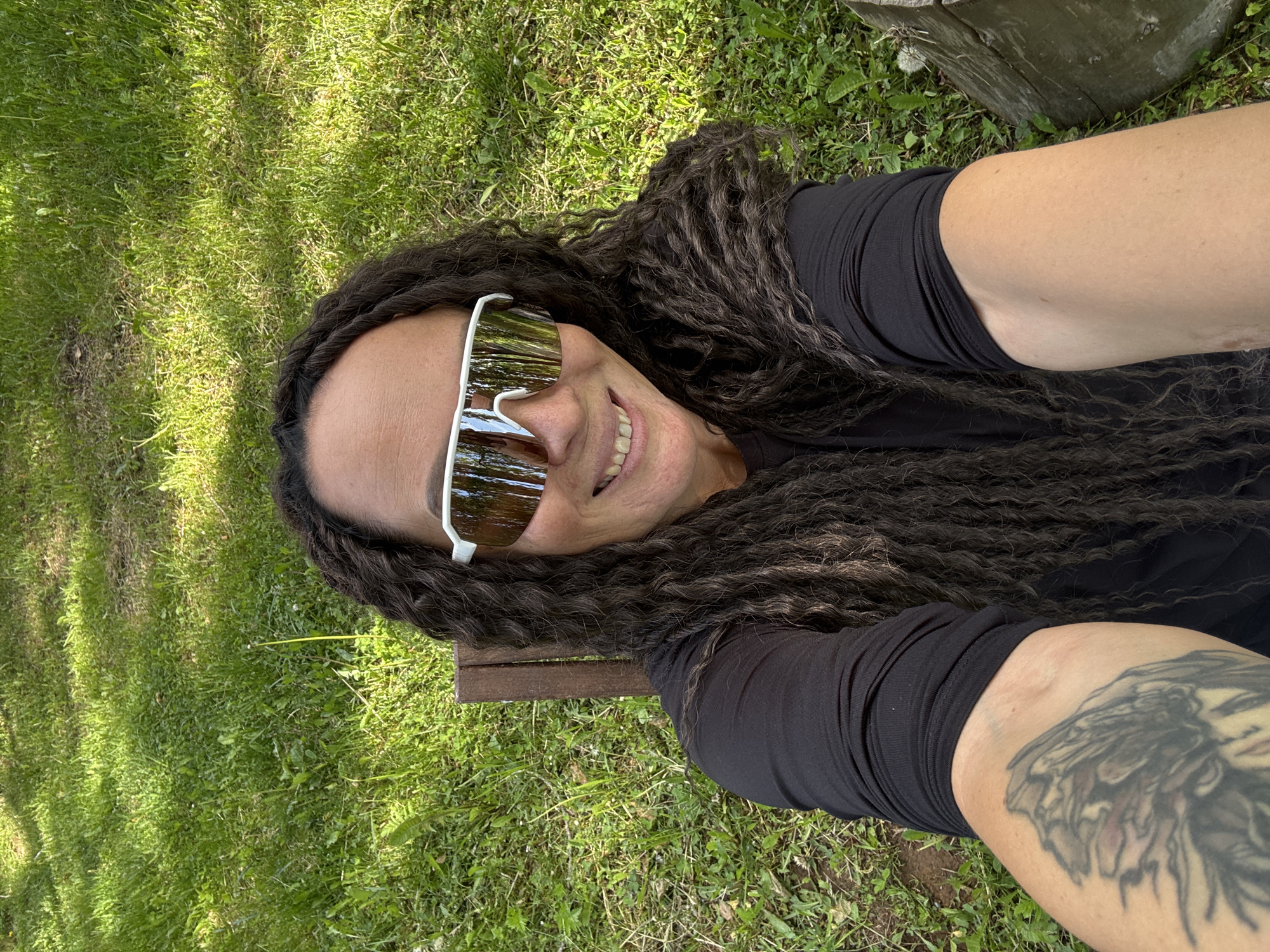 A person with long dark dreadlocks and white-framed sunglasses lies on a patch of green grass. The person is wearing a black long-sleeved shirt. The right arm is extended towards the camera and features a large tattoo. There's a small wooden stool or bench section visible near the person's left side. A partially visible wooden object, possibly a log or piece of furniture, is in the upper right corner of the image. The location is potentially in Chaikovsky, Russia, although there is no definitive evidence of this within the image itself. The person is smiling slightly.