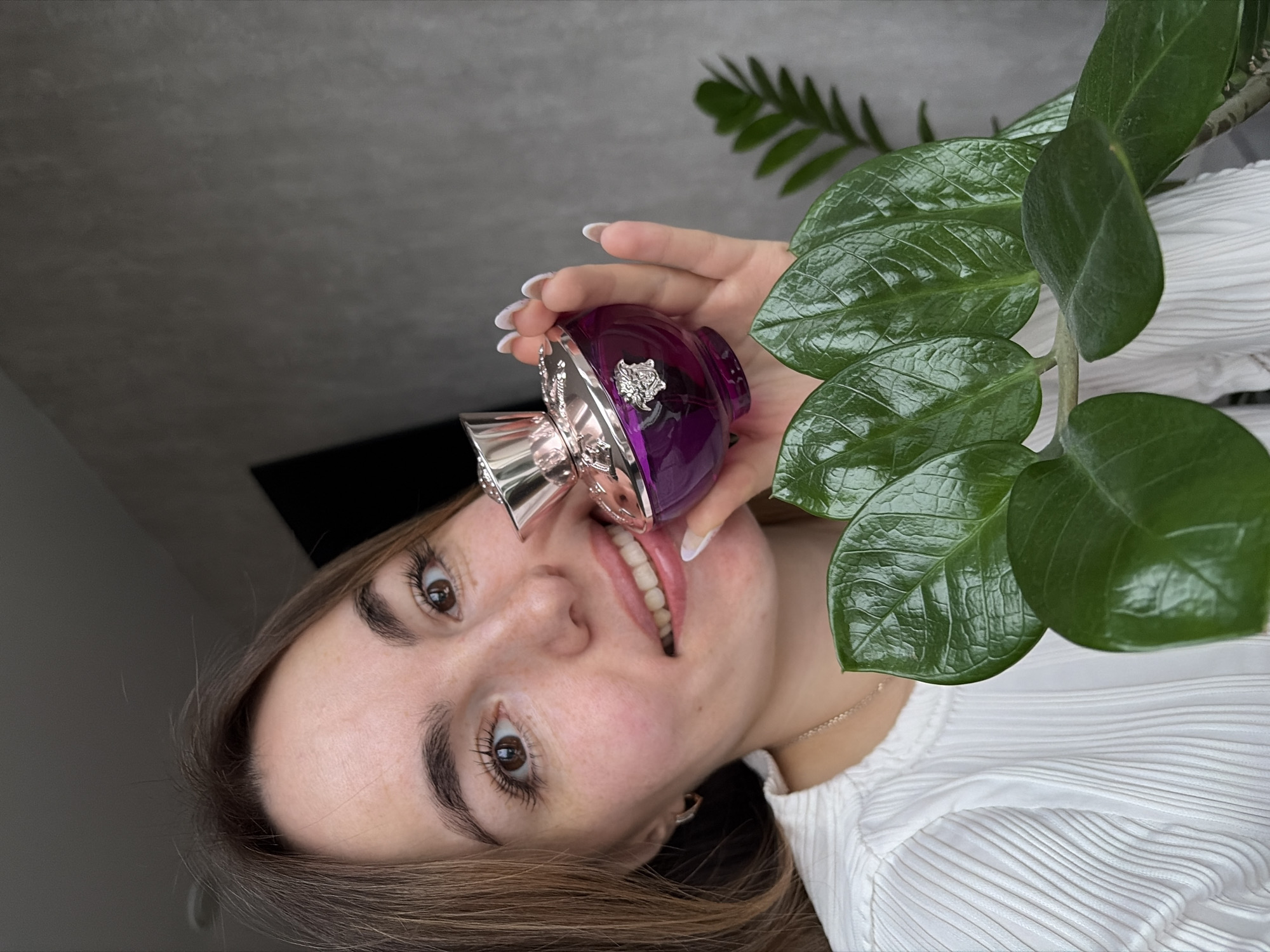 A woman is posing indoors with a bottle of purple perfume. She is holding the bottle near her face with her right hand. She has long brown hair, brown eyes, and manicured nails. She is wearing a white pleated shirt. To her right is a green plant with glossy leaves. The background is a light gray wall. There is no evidence to suggest this photo was taken in Izhevsk, Russia.