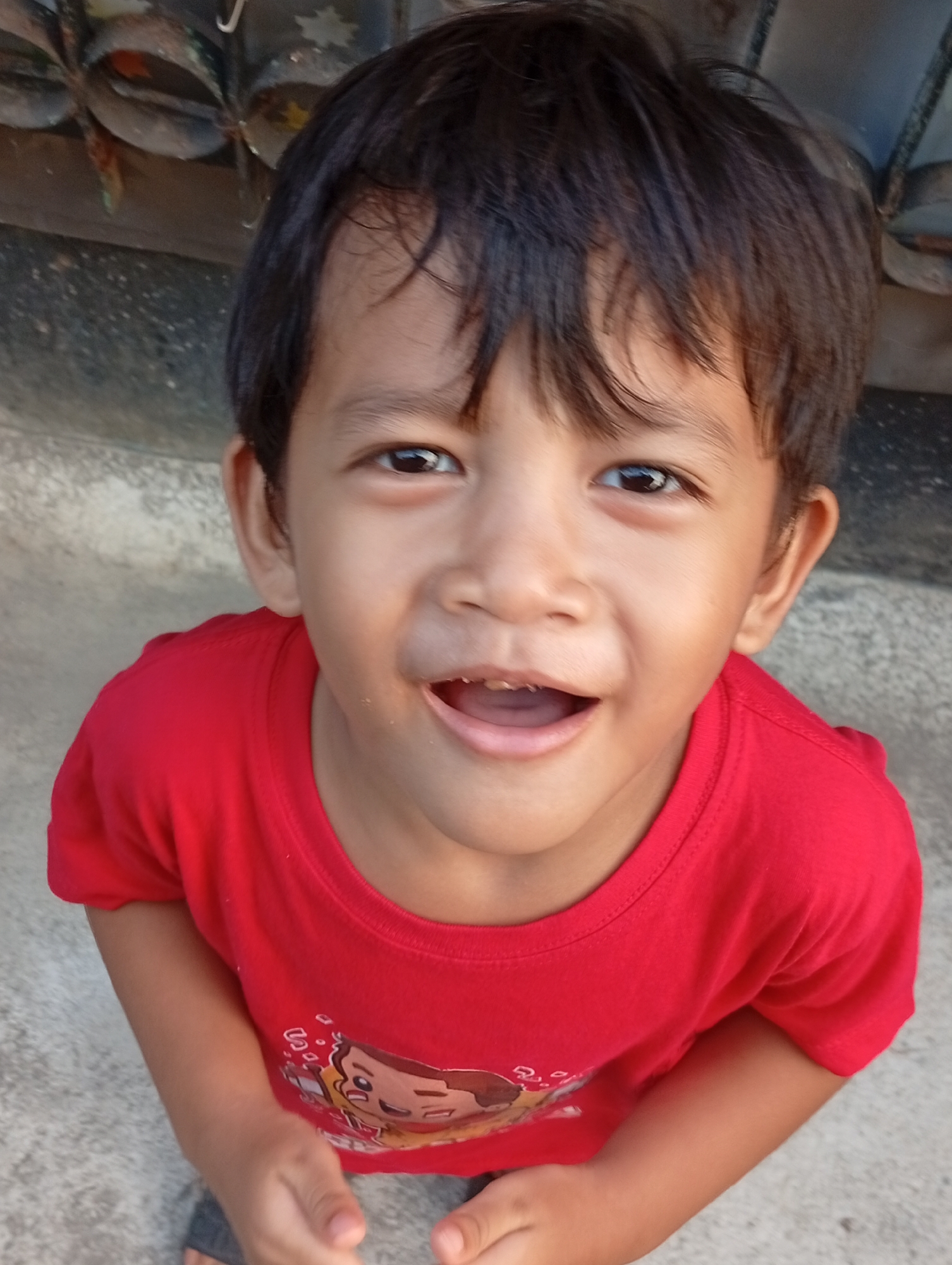 A young Indonesian boy, with dark hair and brown eyes, is captured in a close-up portrait. He is wearing a bright red t-shirt with a graphic print of animated characters. The boy is looking up and smiling with his mouth open, revealing his teeth. His skin tone is medium-brown. The background is out of focus, but appears to be an outdoor setting with a textured concrete surface and what looks like a metal fence or structure above. The lighting suggests it is daytime. This image was taken in the city of Bekasi, Indonesia.