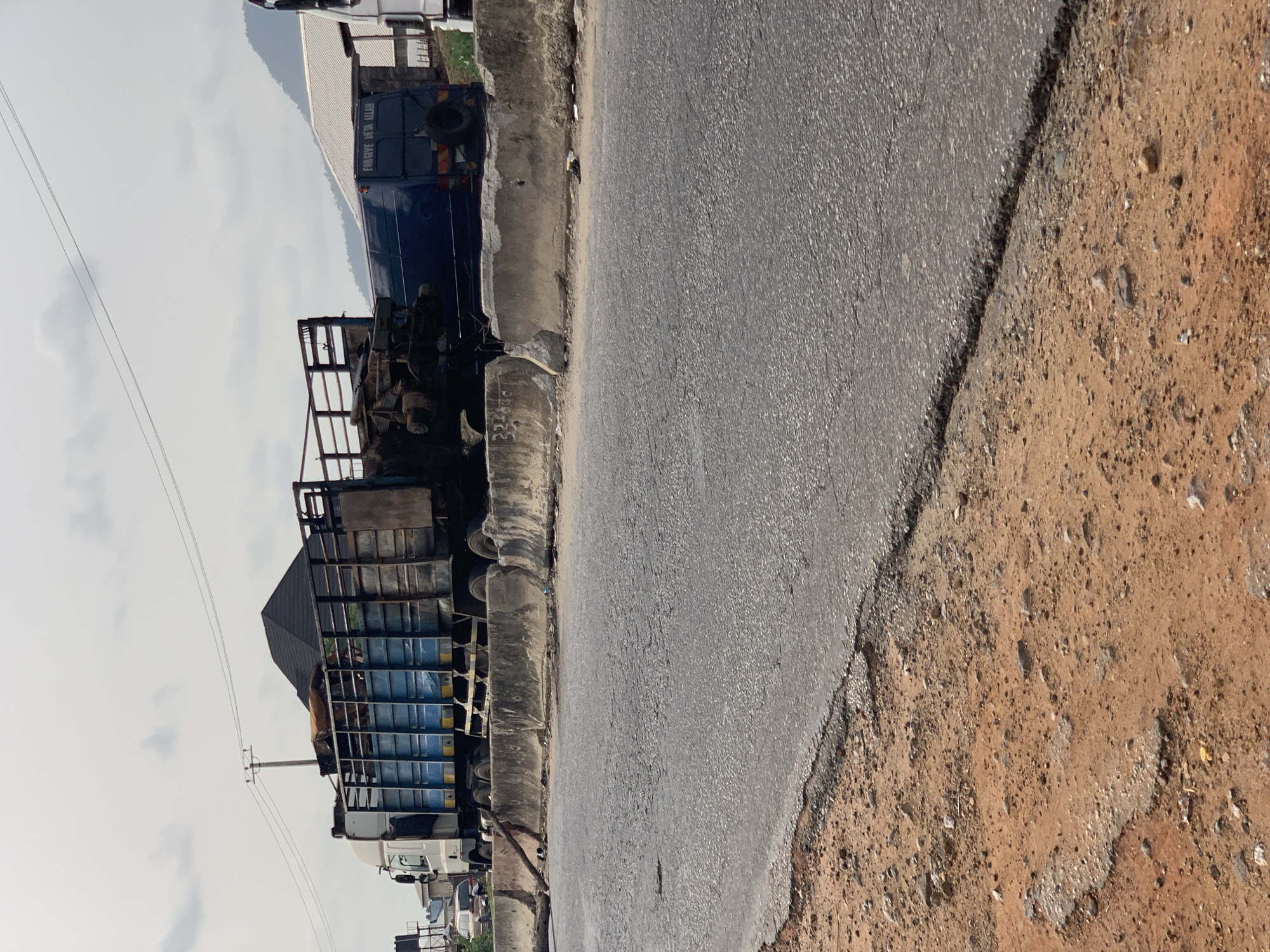 This daytime photograph, taken in Lokoja, Nigeria, captures a roadside scene under an overcast sky. The image is composed with an asphalt road occupying the left side of the frame, leading into the distance. To the right of the paved road is a concrete curb, followed by a wide, unpaved dirt and gravel shoulder.
Along the left side of the road, several vehicles are either parked or slowly moving. A prominent blue truck is visible in the mid-ground, featuring a spare tire mounted on its rear. The text "FORGIVE BETA ALLAH" is distinctly written on the back of this blue truck. Further down the road, another truck with a large, open-sided, metal-framed cargo bed is visible, appearing to carry some goods or crates. Other vehicles, including cars and smaller trucks, are partially discernible in the background. Behind the vehicles, several buildings with corrugated metal roofs are present, blending into the scene. Overhead utility wires extend across the sky, supported by poles. No specific activity is clearly depicted; the scene appears to be a static snapshot of a functional roadway.