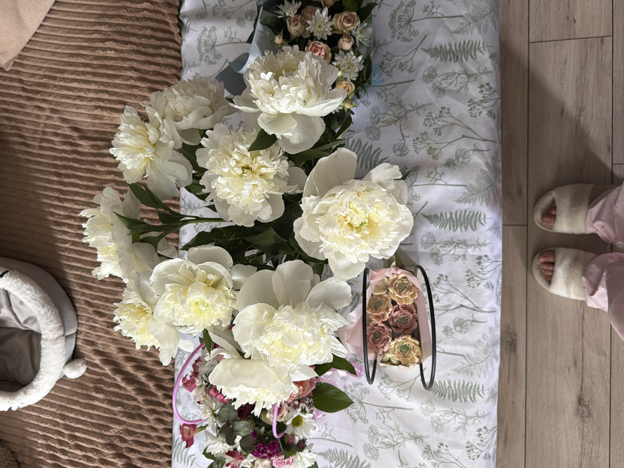 The photograph depicts an aerial view of a bedroom scene. A bed with a white floral patterned sheet is centrally located. A large bouquet of white peonies takes up a significant portion of the frame, arranged on the bed alongside a smaller floral arrangement in a black metal container and another bouquet with smaller flowers. Adjacent to the bed, a brown corduroy-textured surface is visible, with a gray pet bed positioned on it. To the right, wooden flooring extends, upon which a pair of feet clad in pink pajamas and white, fur-lined slippers are situated. The location is specified as Unknown, Unknown.