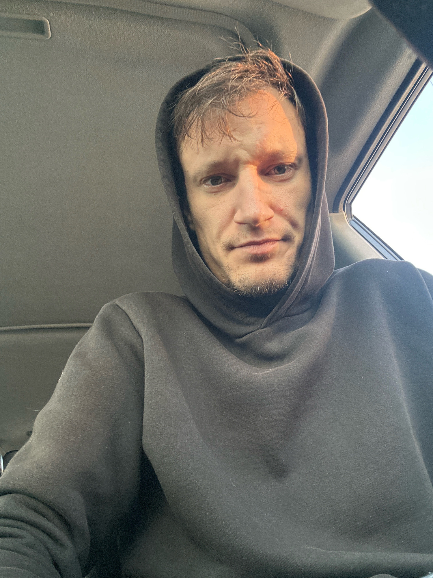 A man, with fair skin, brown hair, and a short beard, is captured in a close-up, selfie-style shot from inside a vehicle. He is wearing a dark hooded sweatshirt, and the hood is pulled up around his head. Sunlight casts a warm glow across the right side of his face, highlighting his features. The interior of the vehicle, including the ceiling and the window frame, is visible. The image is taken in Makiv, Ukraine.