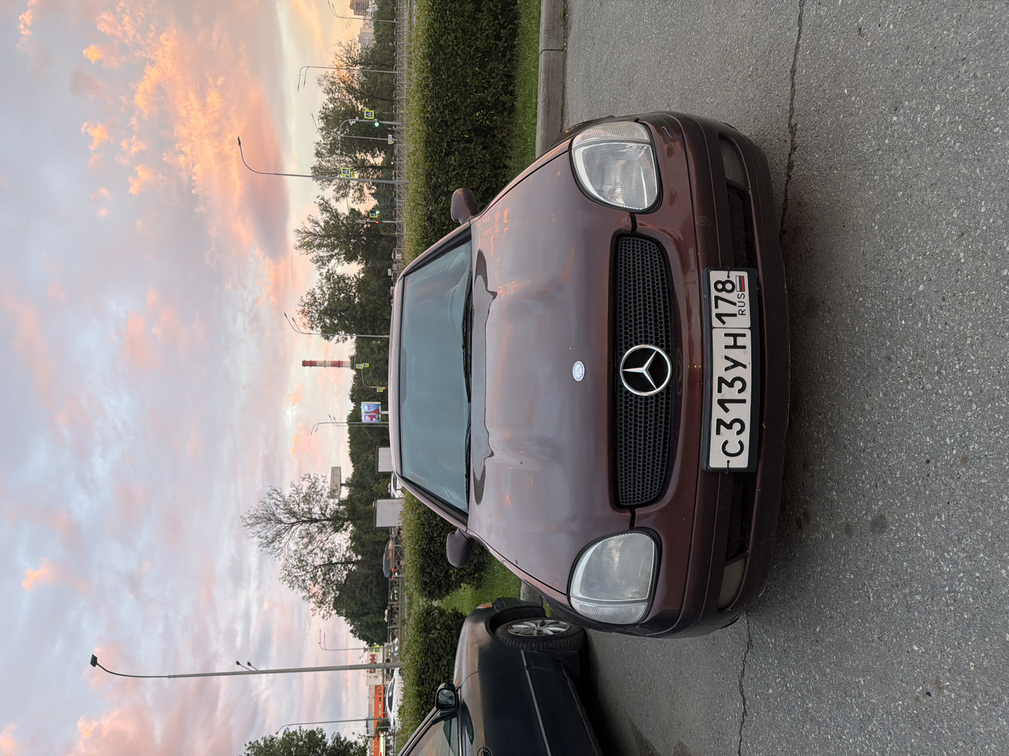 The image shows a front view of a maroon Mercedes-Benz SLK-Class R170 parked on asphalt. The car has a black grille with a silver Mercedes-Benz logo. A license plate with the characters "c313YH 178 RUS" is visible. The car is positioned next to another dark-colored car, partially visible on the left. In the background, there are green bushes, streetlights, traffic lights, trees, and a cloudy sky with pink and orange hues, indicating dusk or dawn. A factory chimney is visible in the far background.
