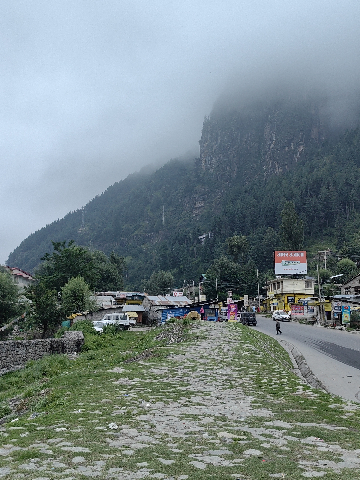 The image depicts a street scene in Bahang, India, with a backdrop of a dense forest covering a steep mountainside. The mountain is partially obscured by fog and clouds, lending a dramatic and somewhat muted atmosphere to the scene. In the foreground, a paved walkway made of irregular stones and patches of grass leads towards the street. To the left of the walkway is a low stone wall.
Along the right side of the street, a row of shops and buildings are situated. These structures appear to be constructed from corrugated metal and stone, with some displaying colorful signage. One prominent billboard features Hindi text and the website address "amarujala.com". Several vehicles are visible on the street, including a white car, a black SUV, and what appears to be another white vehicle partially hidden behind some buildings. A few individuals are present: one person is walking on the street, and another can be seen near the buildings on the left. The overall environment suggests a mountainous region with a developed commercial area along the roadside. Electrical poles and wires are visible, indicating the presence of infrastructure.