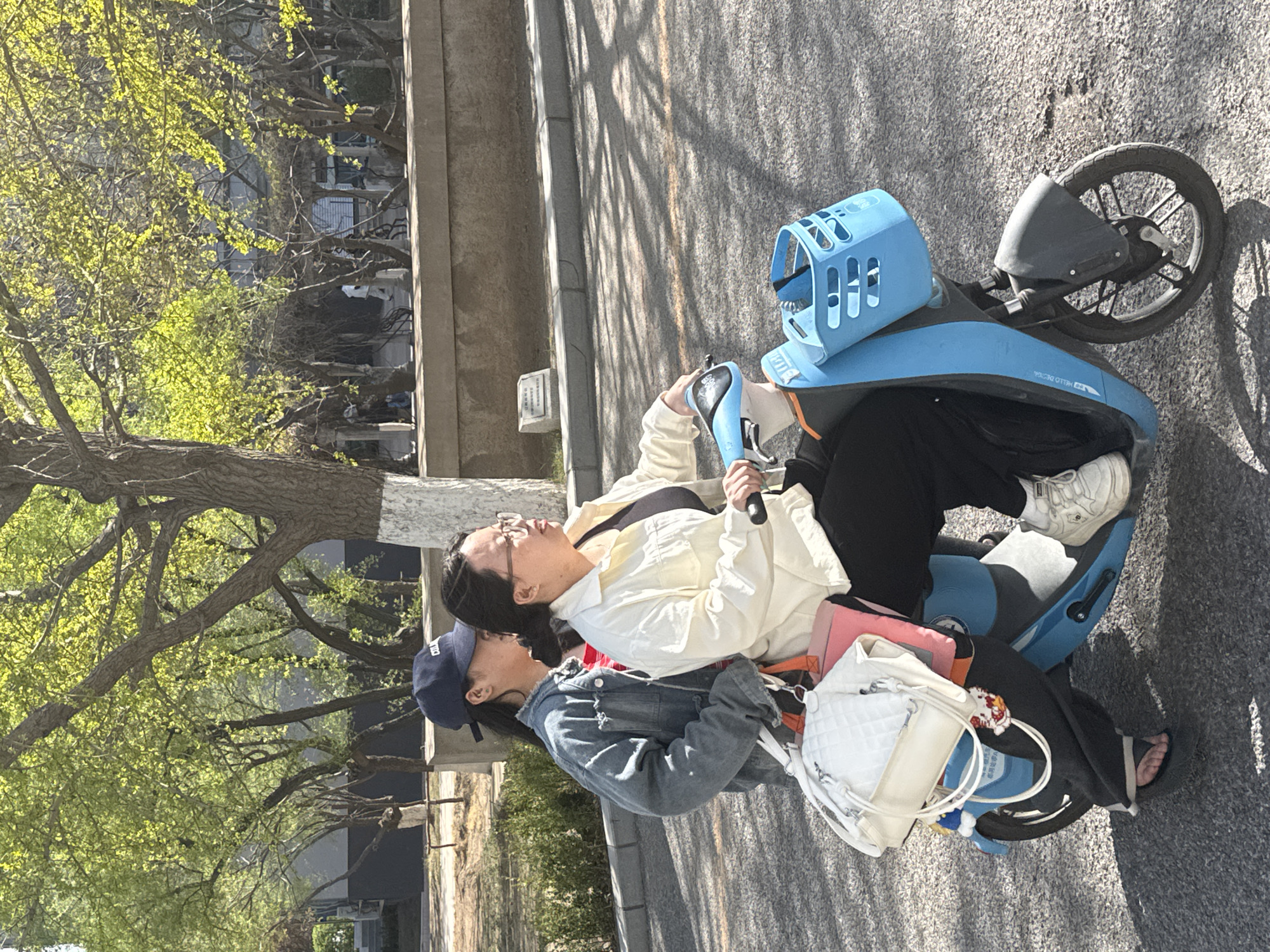 Two women are riding a blue shared e-bike on a paved road. The woman operating the e-bike is wearing a white button-down shirt, black pants, and white sneakers, while the passenger is wearing a denim jacket and a blue baseball cap. A white handbag and a pink object are placed on the seat between them. The background shows a street lined with trees, and a building. The location is in the city of Zhongtun, China.
