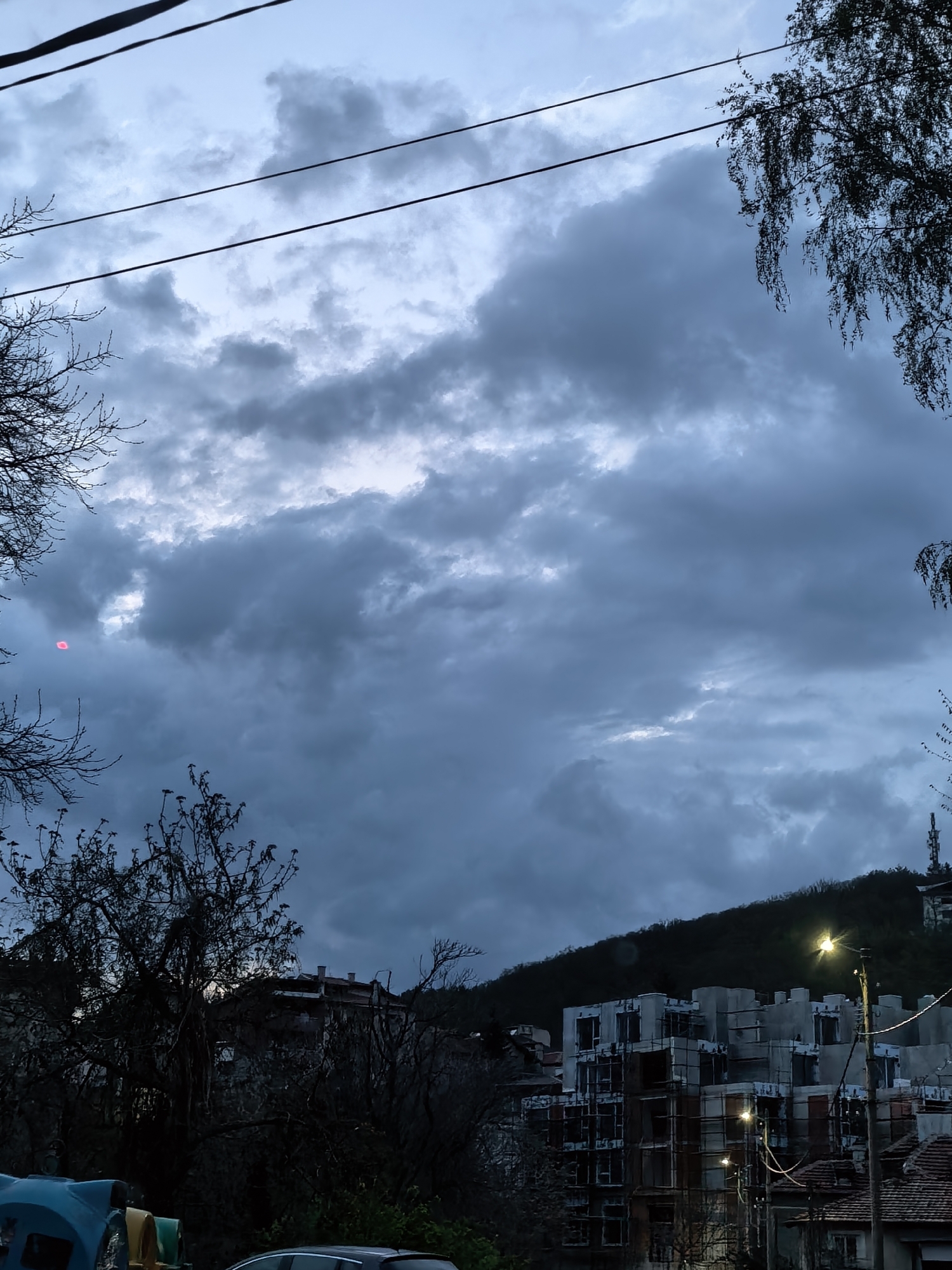 The image captures a cityscape under a cloudy, overcast sky. In the lower part of the frame, there are buildings in various states of construction, with scaffolding and bare concrete visible. Streetlights illuminate the structures. Above the buildings, a tree-covered hill rises into the frame. Bare tree branches are visible in the foreground, framing the scene on both sides. Two electrical wires stretch across the upper portion of the sky. A red dot or lens flare is visible on the left.