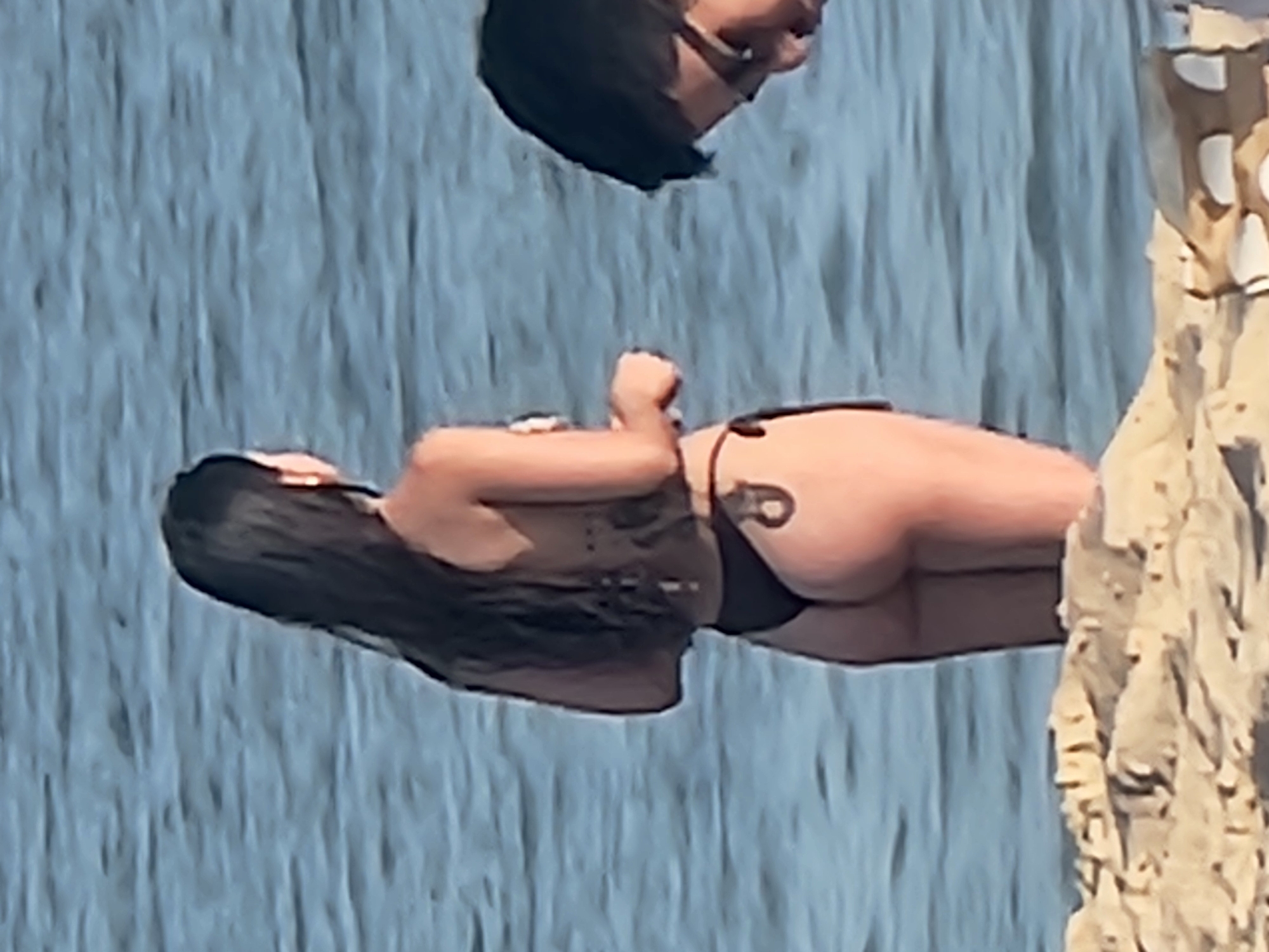 An image taken in Quarteira, Portugal shows a woman from behind, wearing a dark bikini, standing in the ocean. Her dark hair is long and flows down her back. To the right, a person's head with dark hair and glasses is visible, facing downwards. The background consists of rippling blue water and a sandy foreground.