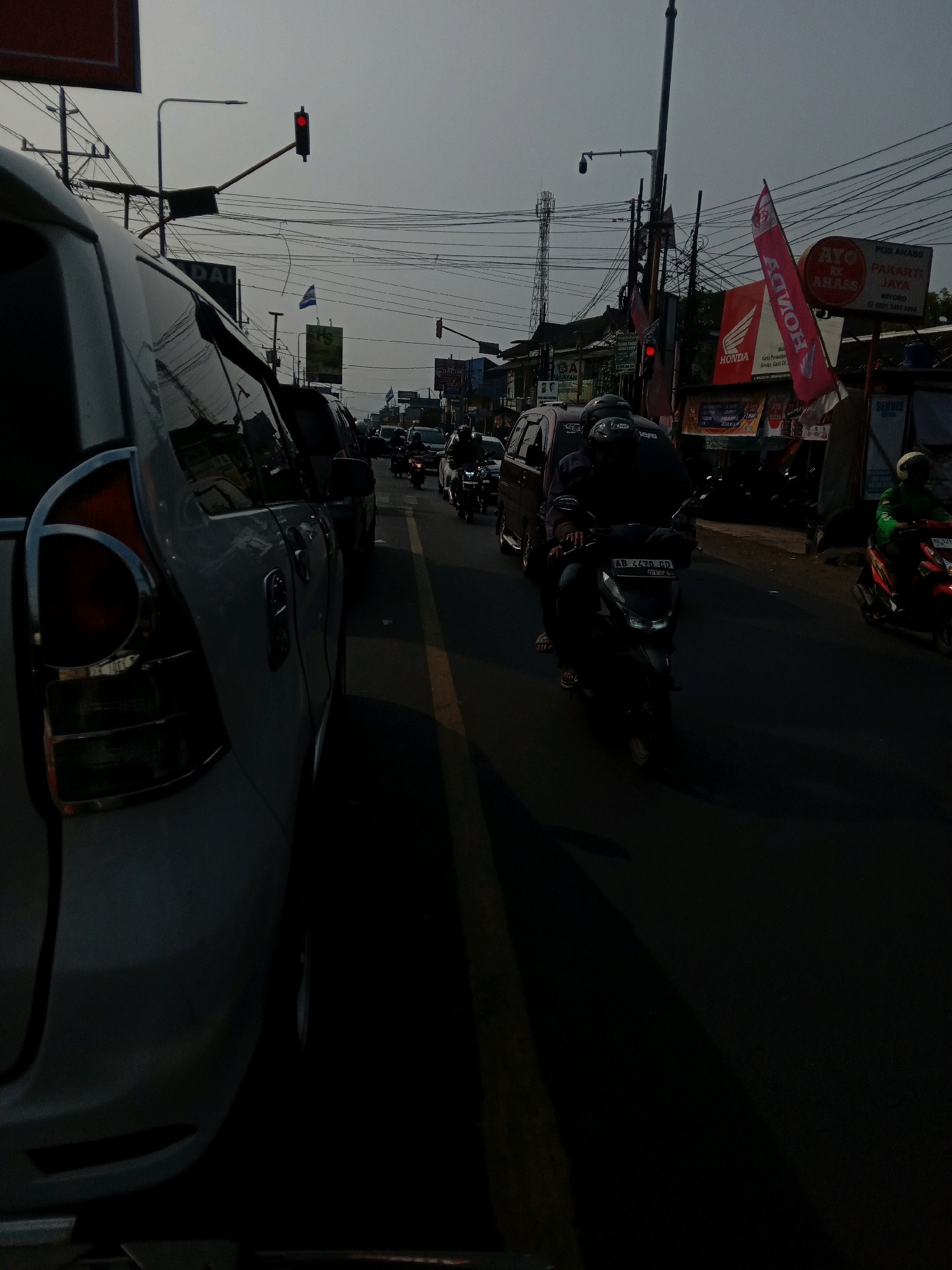 The image captures a street scene in Banguntapan, Indonesia, likely during daytime given the traffic and lighting. In the foreground, the rear of a white SUV is prominently displayed on the left side of the frame, its taillight assembly visible. The vehicle is in what appears to be a traffic lane, indicated by the yellow dividing line on the asphalt road. A dark shadow stretches across the road from the SUV.
Further down the road, traffic is moderate to heavy. Several motorcycles are visible, with riders wearing helmets. One motorcycle in the mid-ground, bearing the license plate AB 4470 GD, is being ridden by two individuals. To the right, another motorcycle with a rider in a green jacket is moving. In the background, there are buildings lining the street, some with signs and flags. Notably, a red traffic light is illuminated, suggesting a controlled intersection. Power lines and poles crisscross the sky, a common sight in urban Indonesian landscapes. Various advertisements and signs, including one that reads "AYO PAKARTI JAYA," are affixed to the buildings. The overall atmosphere suggests a typical bustling street in an Indonesian city.