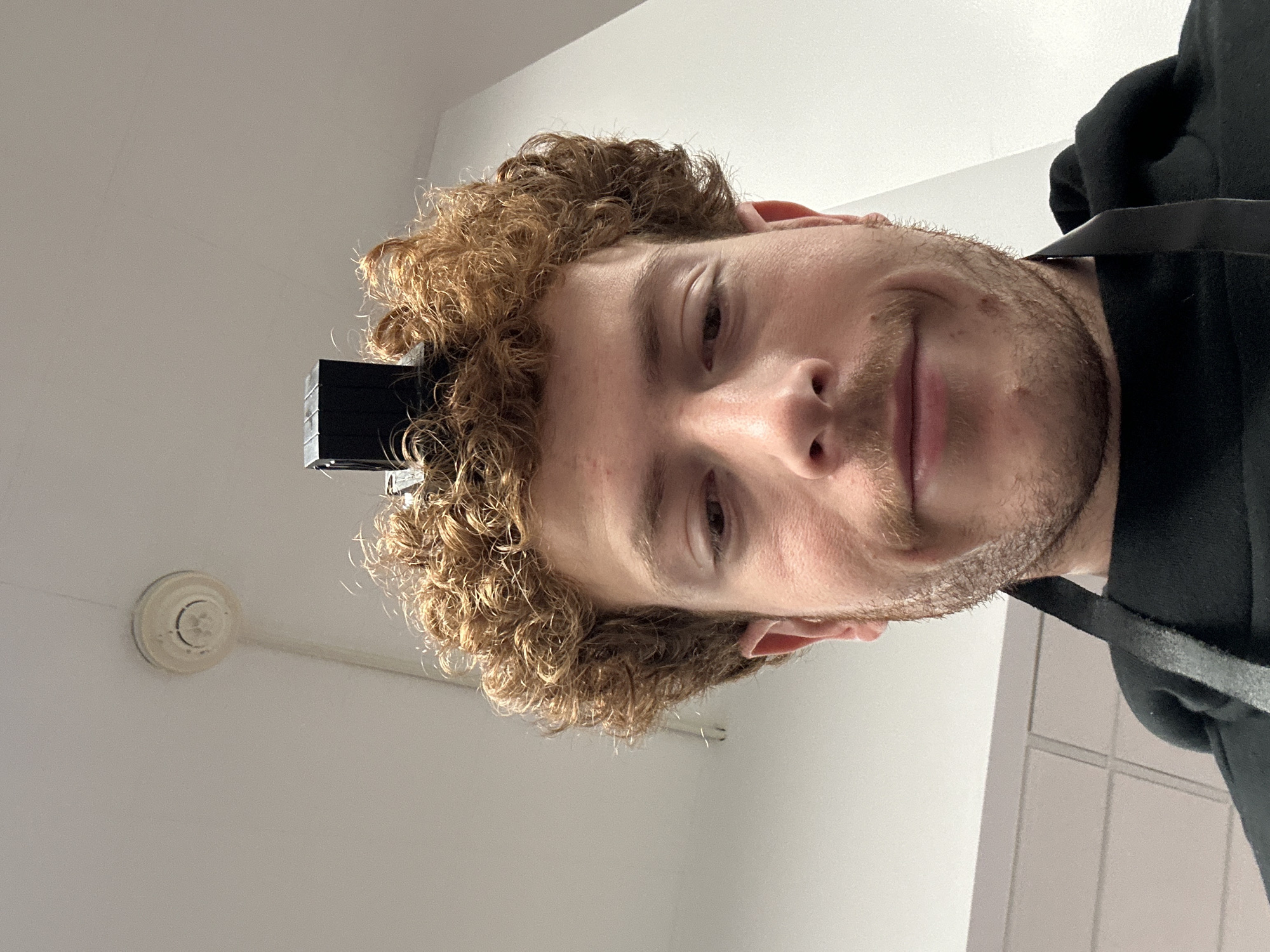 An image taken in an interior setting in the city of New York, United States of America, depicts a young adult male from a low-angle perspective, looking directly into the camera. The male has reddish-brown, curly hair, a light brown mustache, and stubble. He wears a black hooded sweatshirt, with a black strap visible over his left shoulder, possibly belonging to an apron or bag. A small, black, rectangular object rests on the crown of his head, partially obscured by his hair, consistent with a tefillin box. The background features a plain white ceiling with a circular white smoke detector or fire alarm mounted in the upper left. A light-colored wall or set of cabinets with rectangular paneling is partially visible in the lower left background.