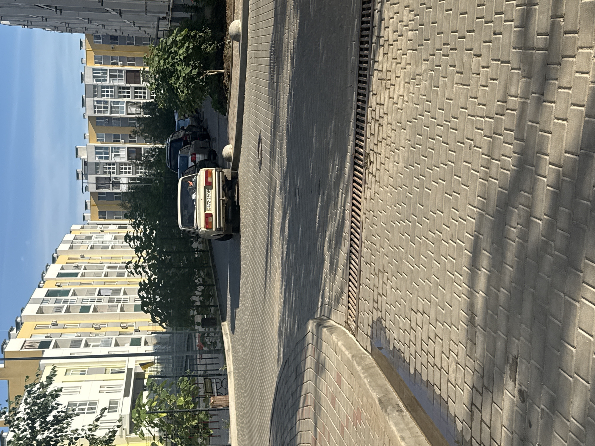 This is a vertical, eye-level, daytime, outdoor photograph taken in Avanhard, Ukraine. The scene depicts a street lined with modern residential buildings and parked cars. The buildings are multi-story structures with light yellow and white facades, featuring numerous windows and balconies. Trees and foliage are visible along the street, casting shadows on the paved walkway.
In the foreground, the walkway is constructed of grey interlocking paving stones. A drainage grate runs parallel to the curb. Several cars are parked along the side of the street, including a white sedan in the center with a visible license plate. The sky is clear and blue, indicating fair weather. The time of day appears to be late morning or early afternoon, given the angle of the shadows.
The overall impression is one of a contemporary urban residential area on a sunny day. There are no people clearly visible in the frame, and the scene appears calm and ordinary. The visible text is on the license plate of the white car, which reads "472-290E".